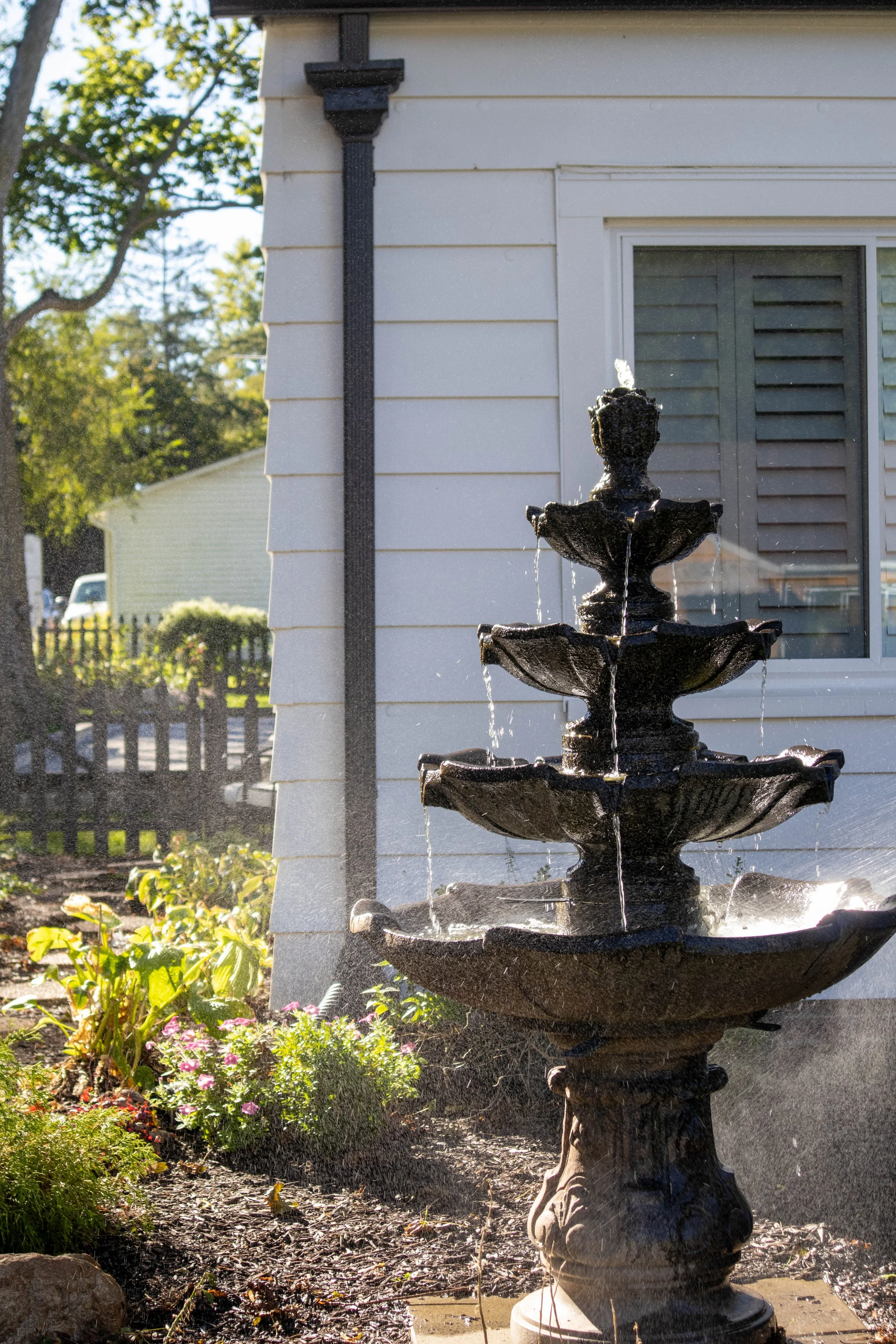 A black, ornate, three-tier fountain with water spouting from the top, located in a garden with green plants and flowers, with a house and trees in the background.