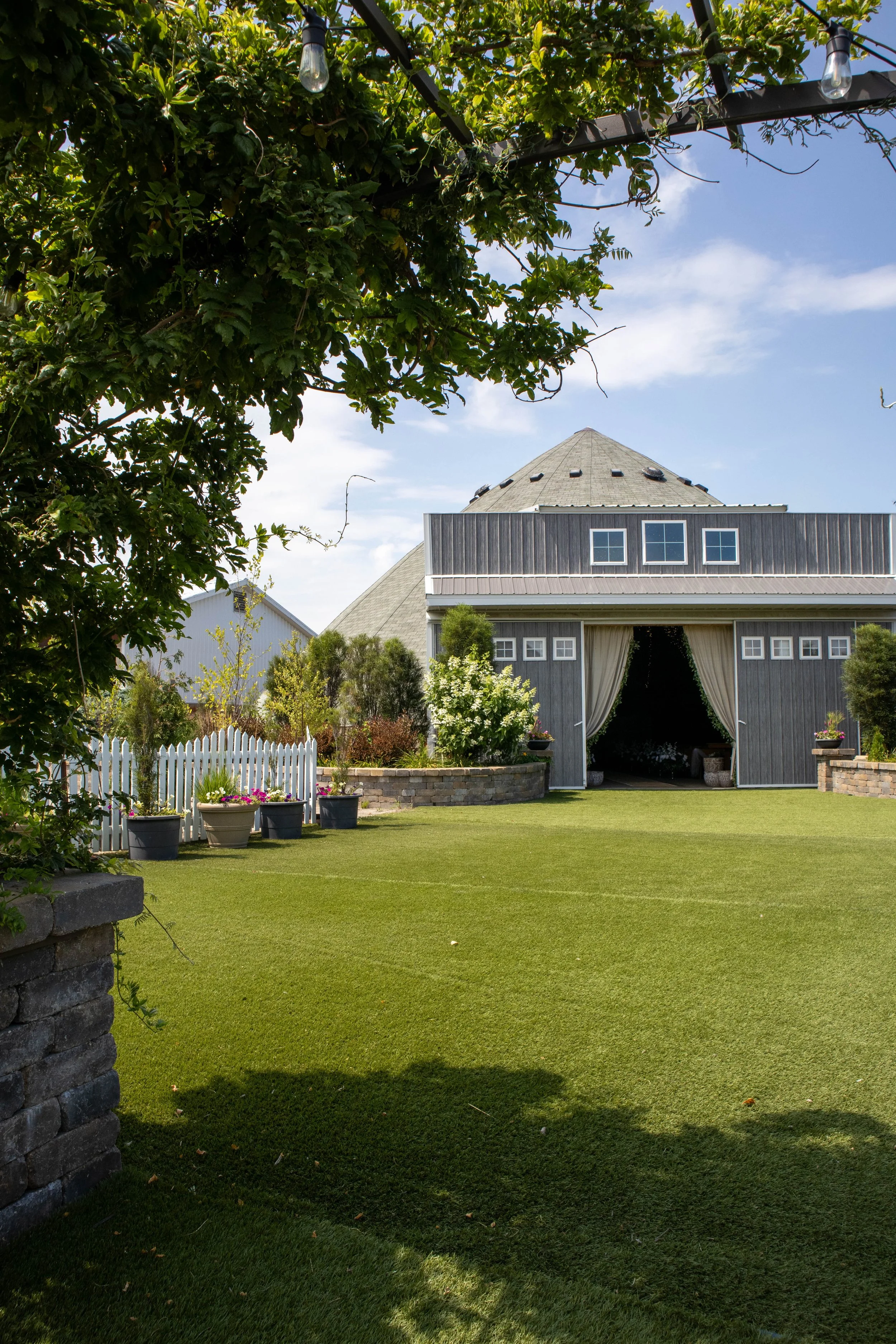 A well-maintained backyard with a large green lawn, potted plants, a white picket fence, and a rustic barn with curtains at the entrance. There are trees and string lights overhead, and a partly cloudy sky in the background.