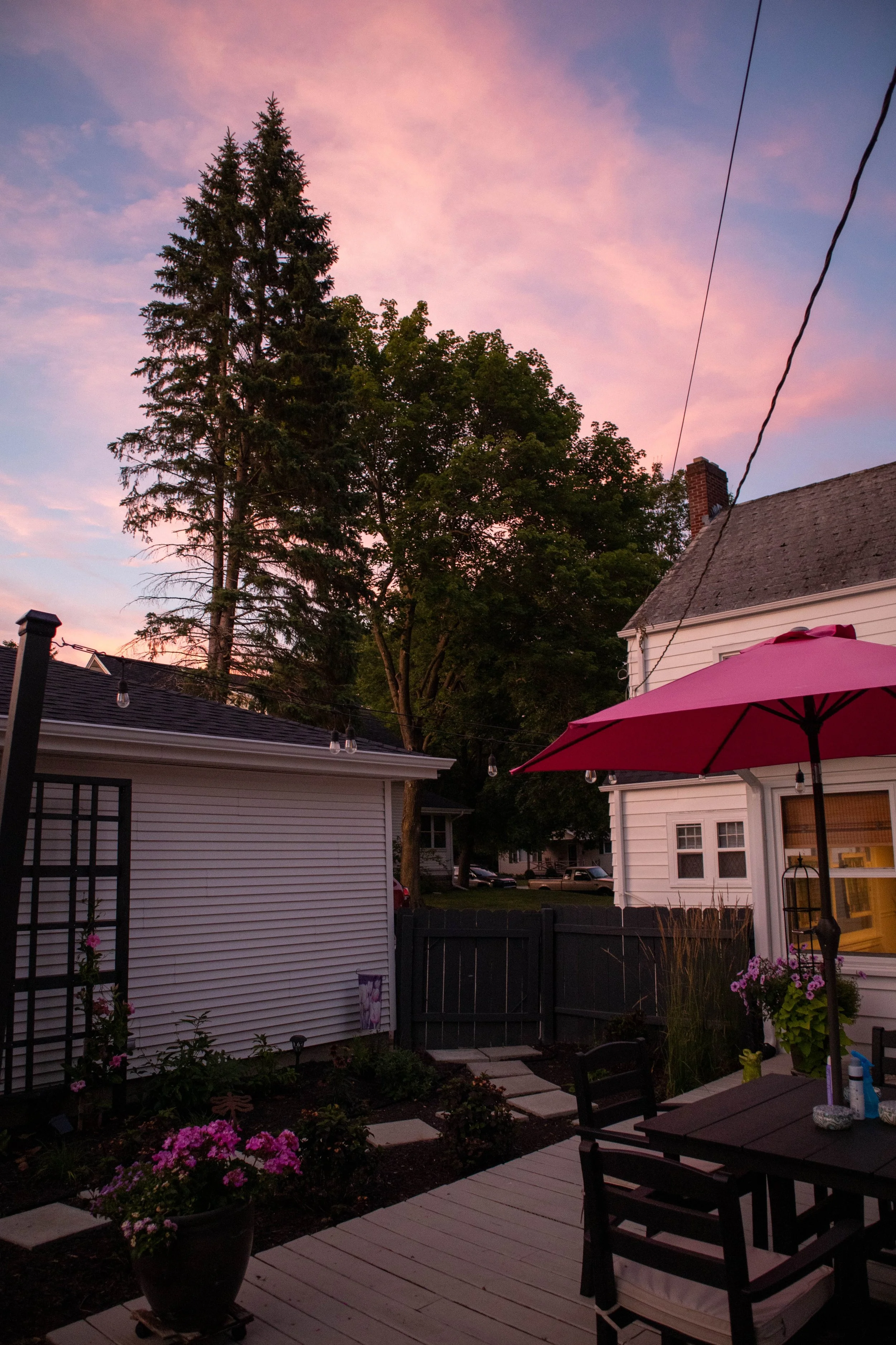 A backyard patio during sunset with pink and purple sky, a pink umbrella over a black table, potted flowers, garden beds, trees, houses, and a fence.