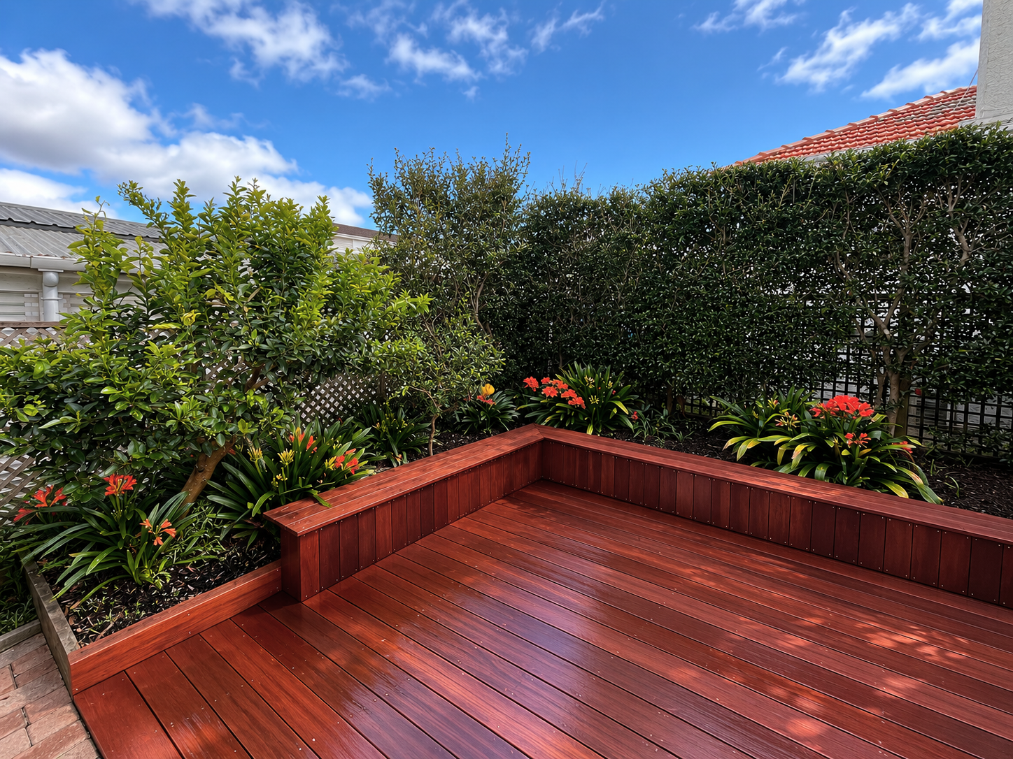 A backyard garden with a wooden deck, lush green bushes, and bright red flowers, under a partly cloudy blue sky.