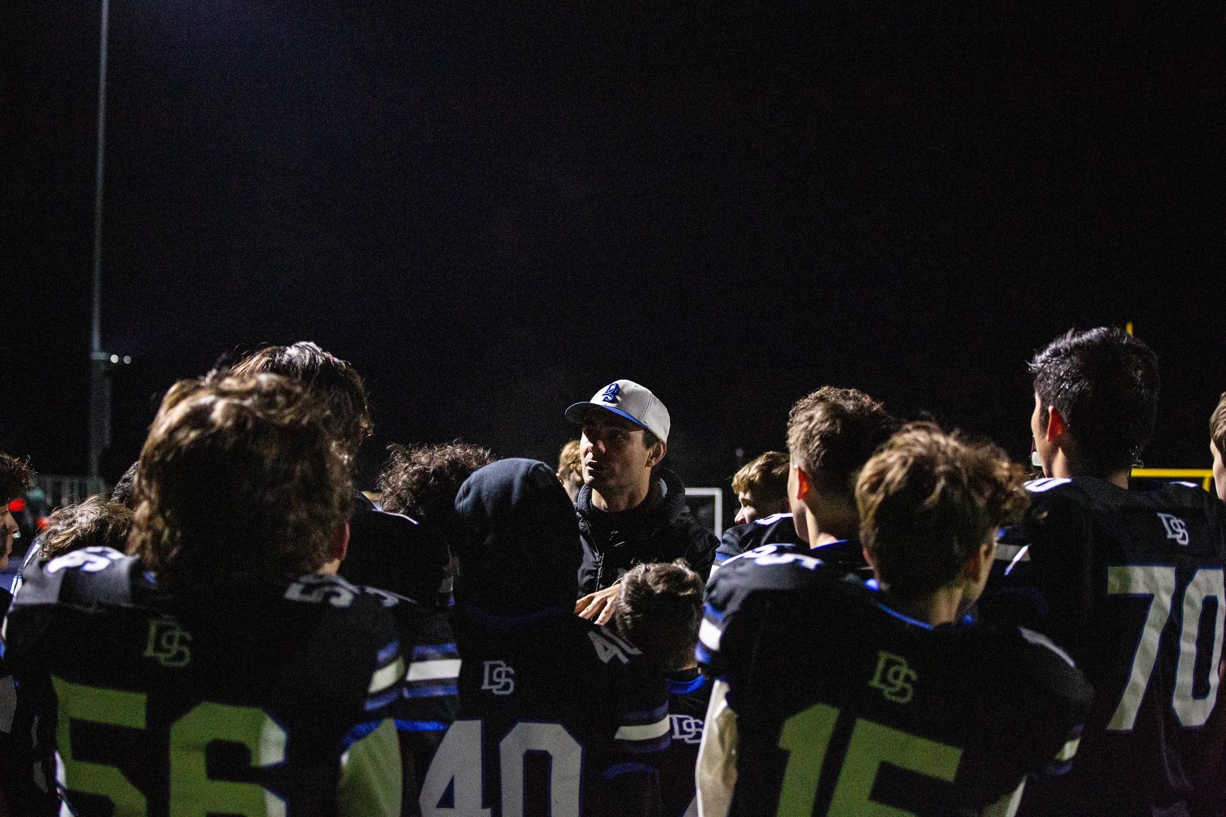 A group of football players and a coach gathered on a field at night, illuminated by stadium lights, wearing uniforms with black, blue, and green accents.