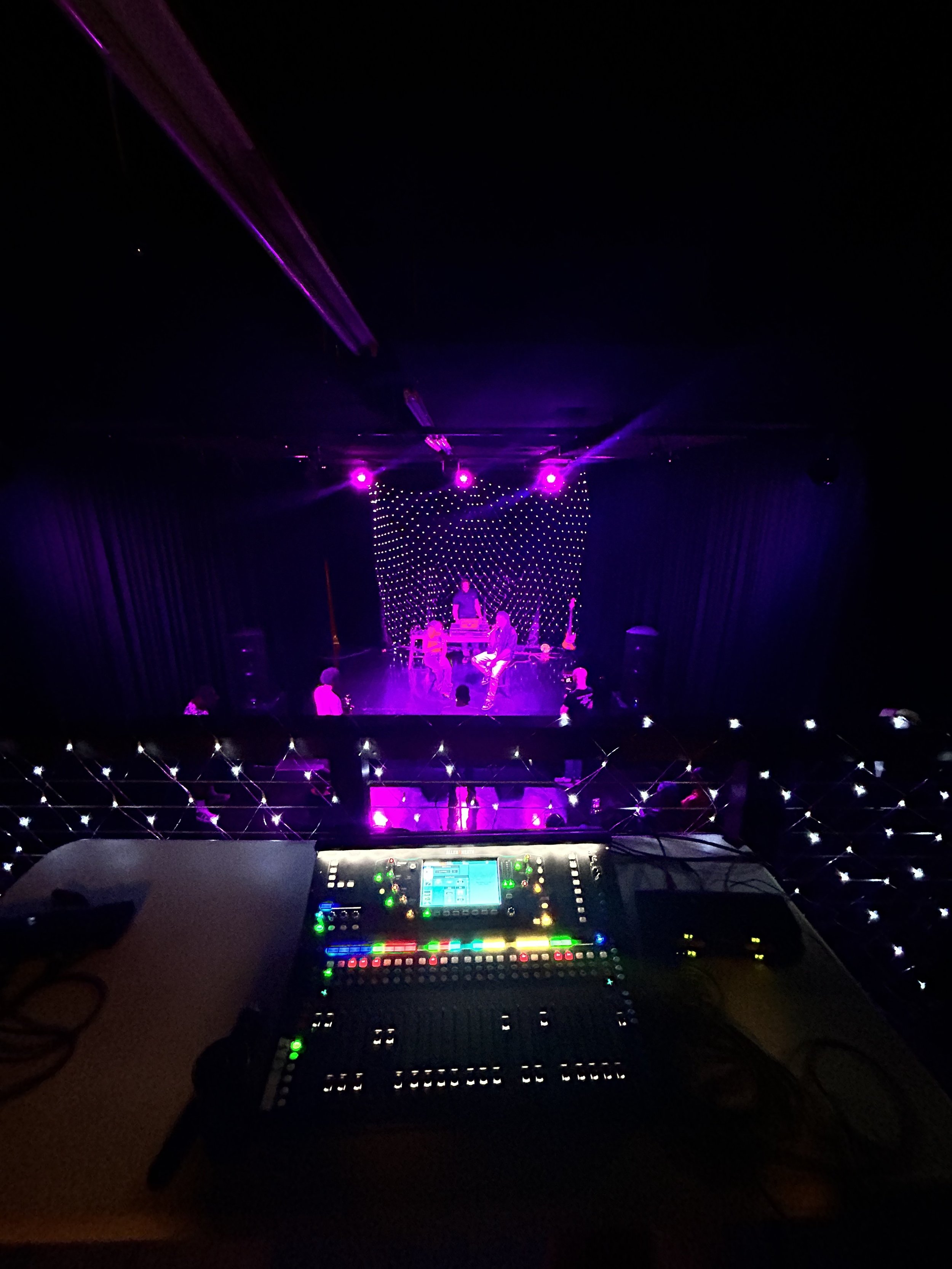Dark stage with purple lighting, DJ booth with equipment in the foreground, and performers dancing on stage with a backdrop of LED lights.