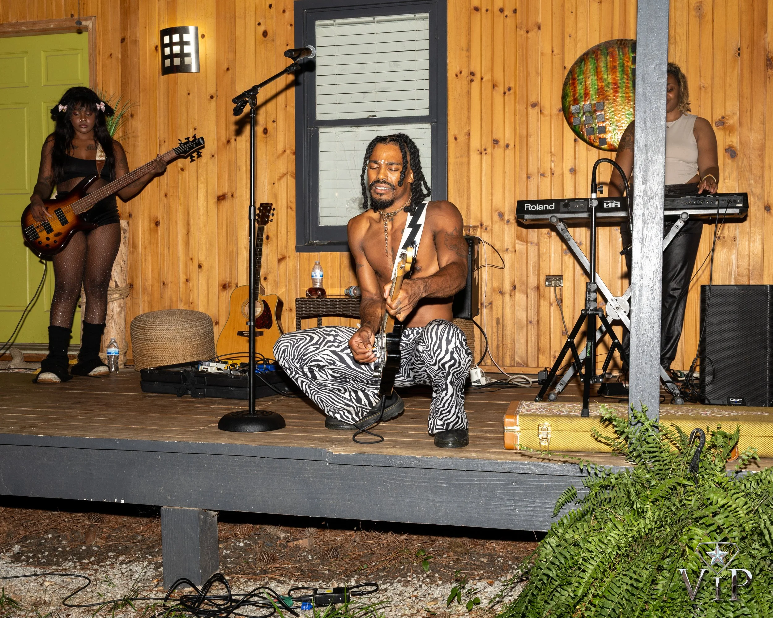 Musicians performing on a wooden stage with a woman playing bass guitar on the left, a man with dreadlocks and zebra-striped pants playing an electric guitar in the center, and a woman on the right using a keyboard. The stage has a water bottle, a small guitar, and various equipment. There are plants in the foreground and a decorative wall hanging behind the keyboard player.