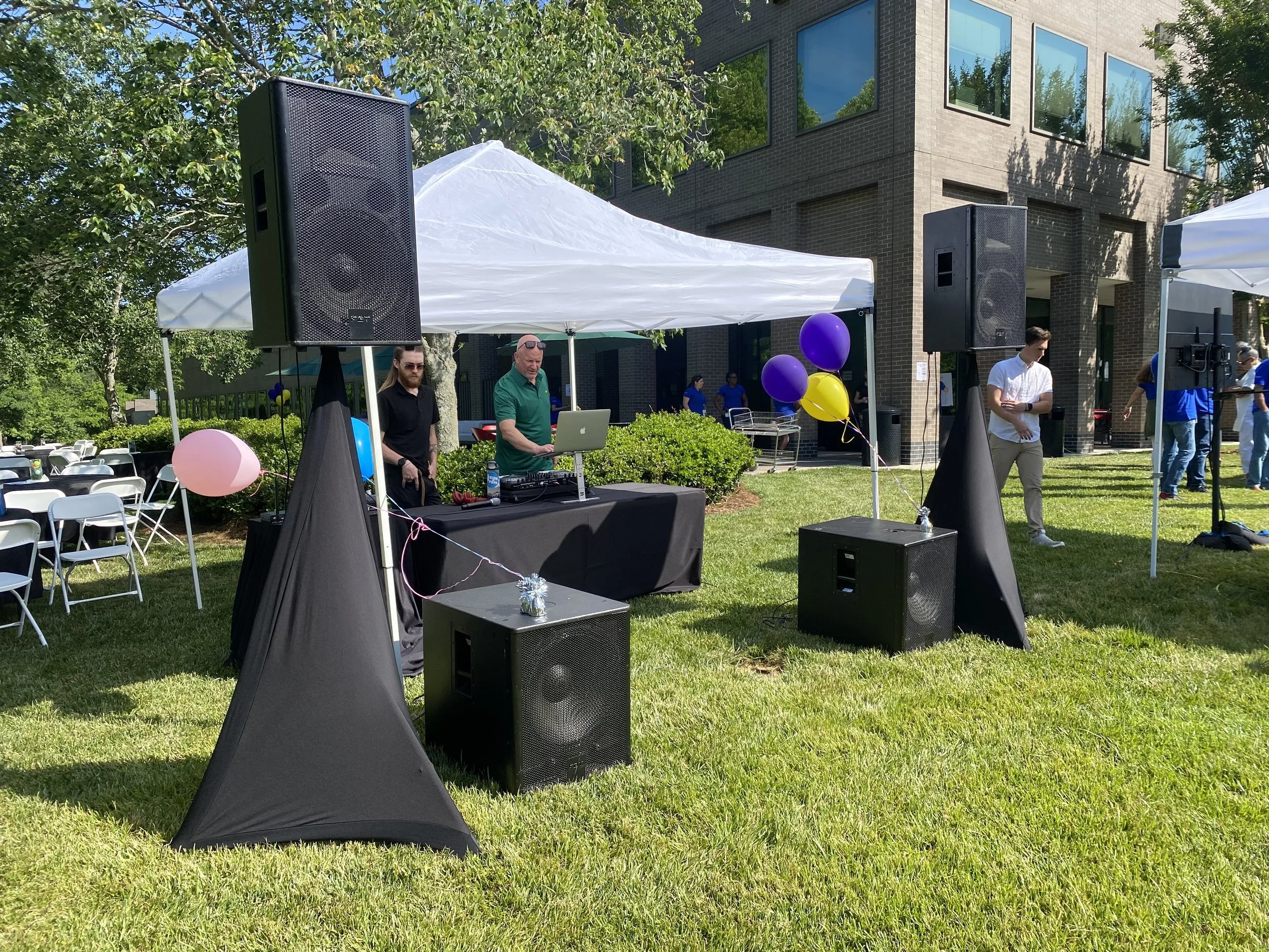 An outdoor event setup with a white tent, large black speakers, balloons, and people, in front of a modern brick building with large windows.