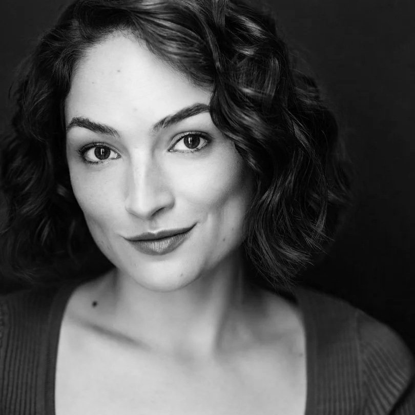 Black and white portrait of a woman with short, wavy hair, looking at the camera with a slight smile, against a dark background.