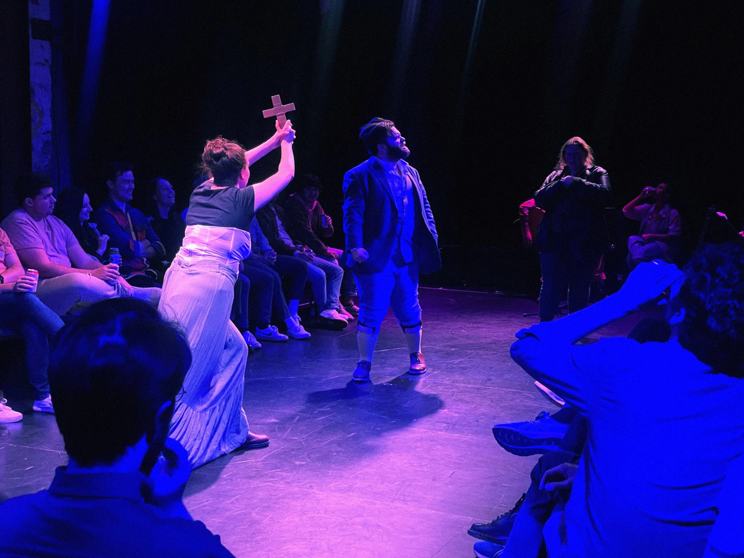 A group of young adults sitting in a dark theater with purple and blue lighting, watching a performance. An actor in the foreground holds up a cross shaped object.