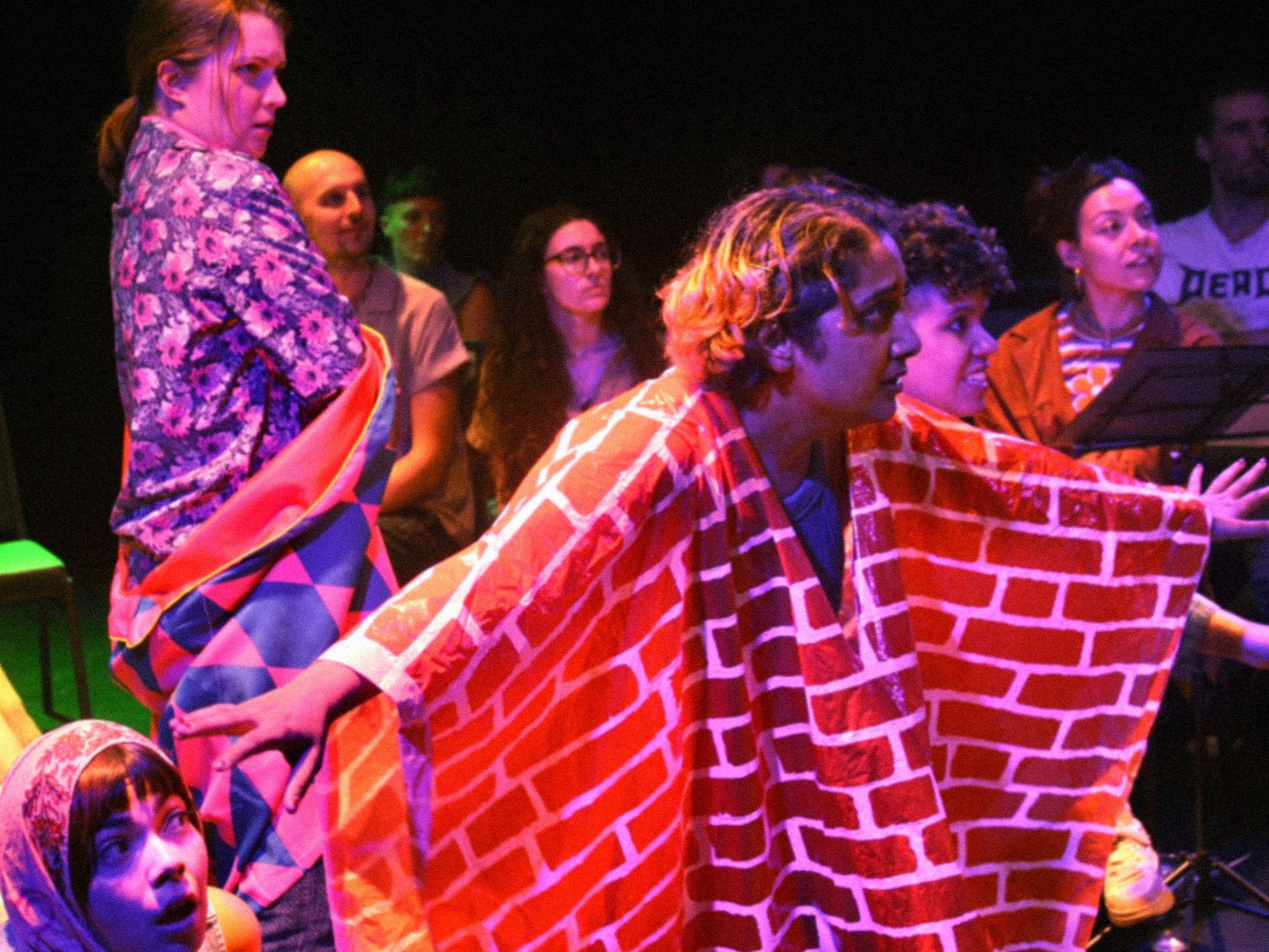 Group of people sitting and standing, one wearing a brick wall patterned costume, in a dark room with vibrant lighting.