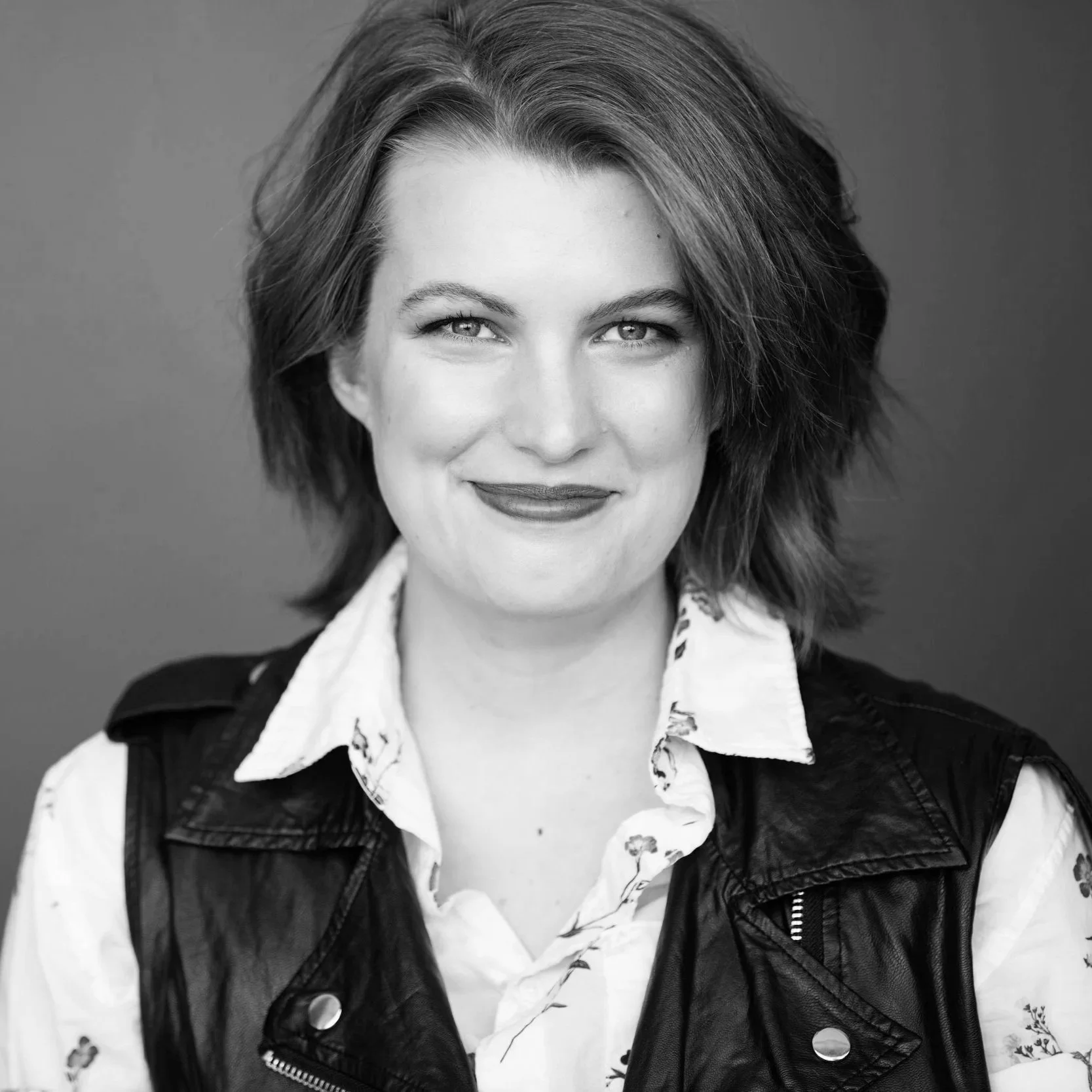 Black and white portrait of a person with short wavy hair, smiling, wearing a floral shirt layered with a leather vest.