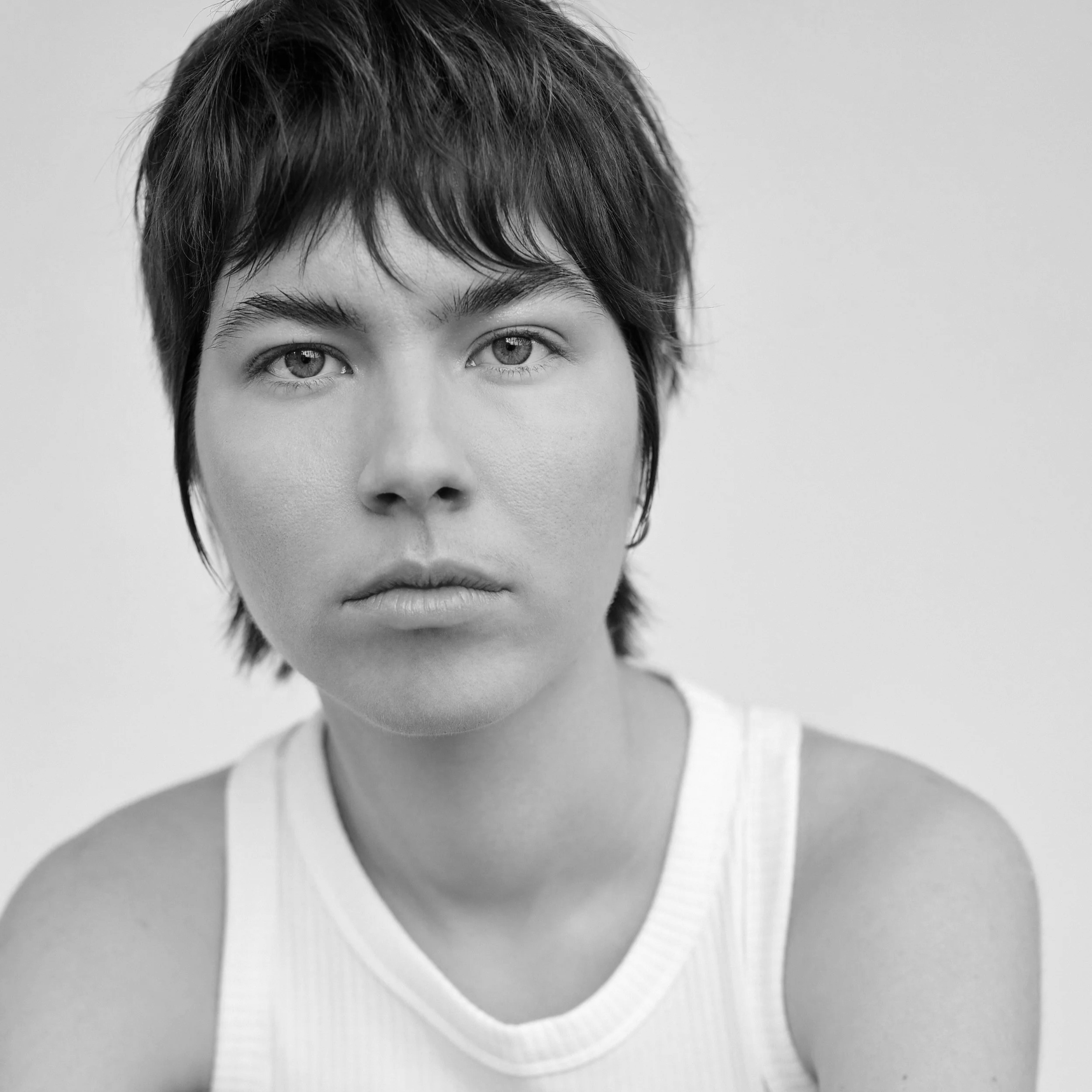 A black and white close-up photograph of a person's face. The individual has short, tousled hair and light-colored eyes. The background is plain and blurred.