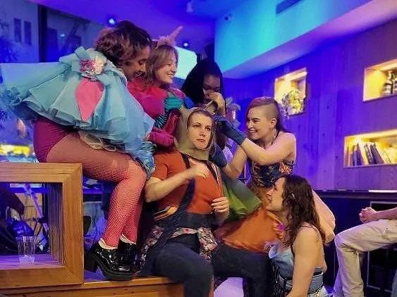 Group of actors in colorful clothing, some with playful accessories, one in a donkey mask, gathered closely in a lively indoor setting with purple and blue lighting, engaging with each other.