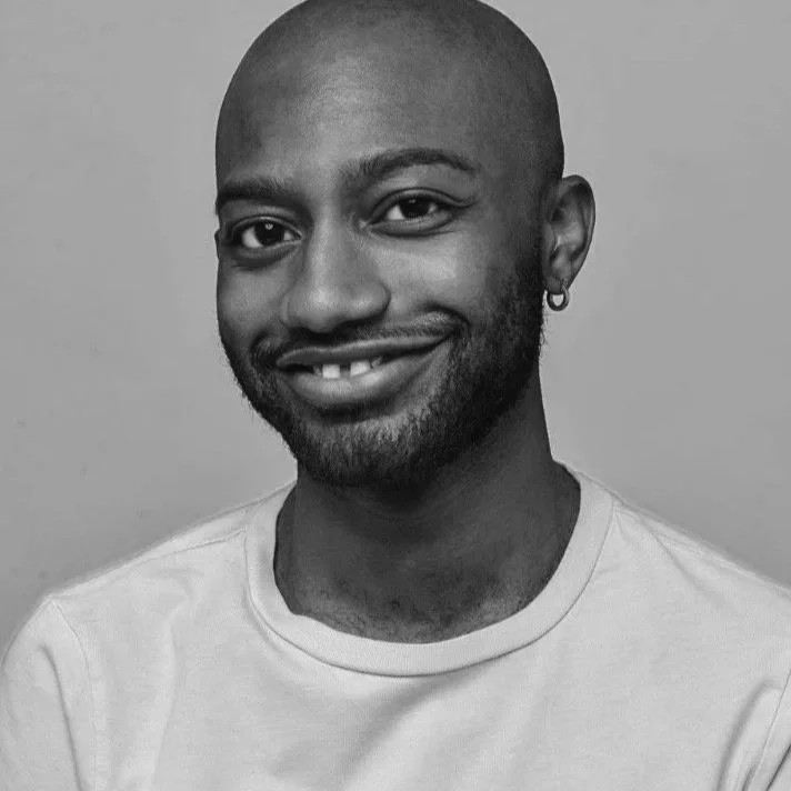 Black and white photo of a smiling man with a short beard, wearing a light-colored T-shirt and a small hoop earring in his left ear, against a plain background.