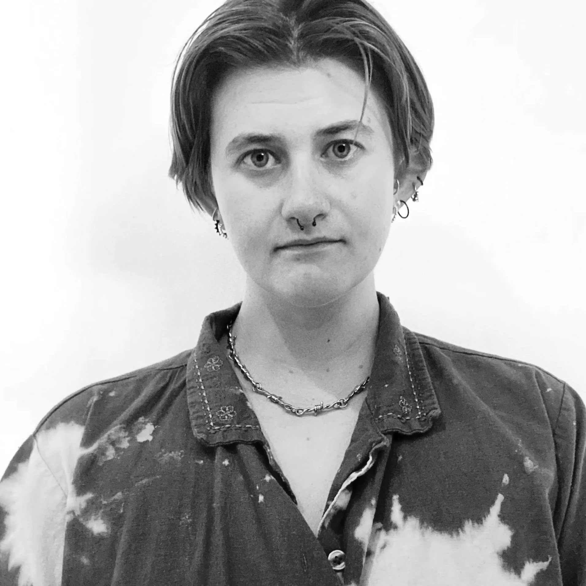 Black and white portrait of a person with short hair, multiple piercings including a septum ring, earrings, and a necklace, wearing a bleached collared shirt, against a plain background.