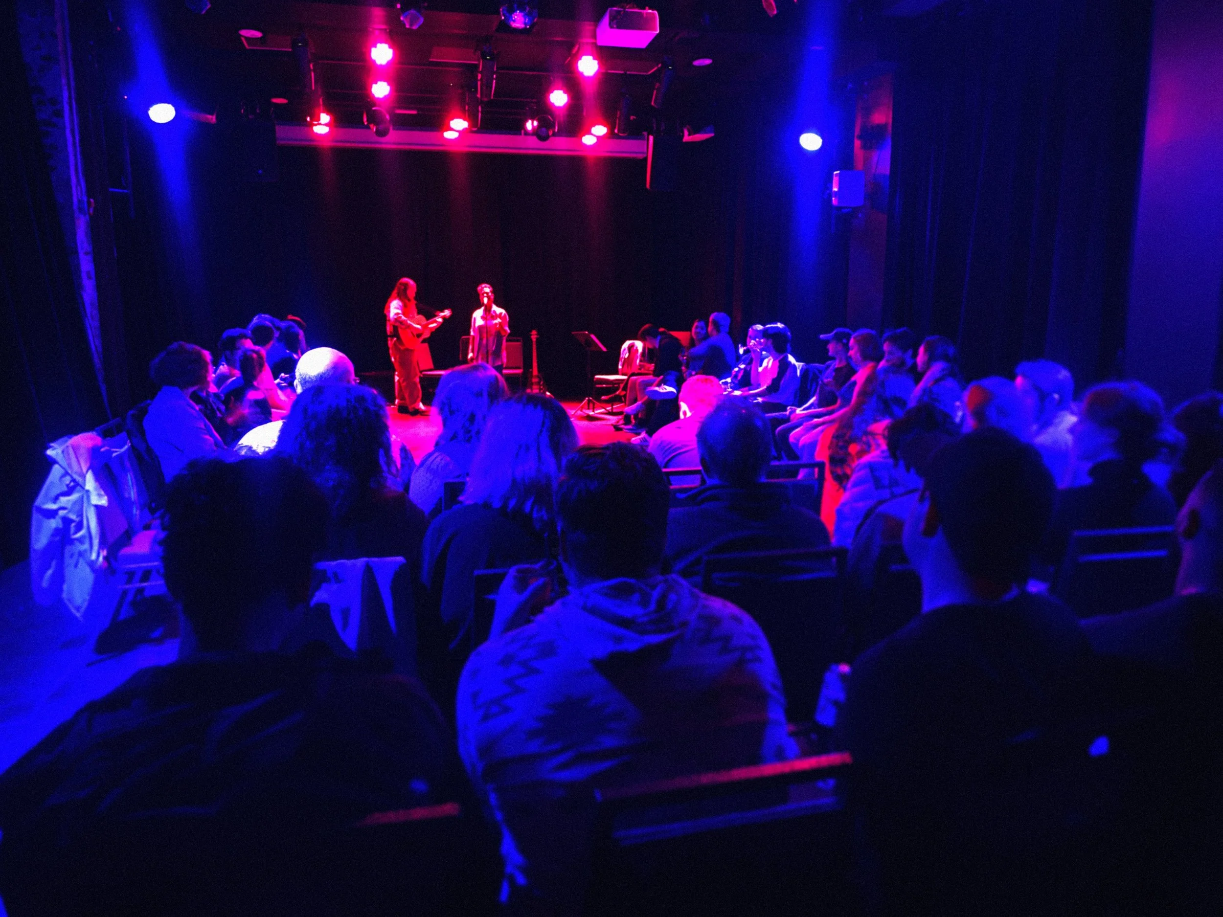 Stage with purple and pink lighting, two performers, one playing guitar and the other singing, audience seated in front.