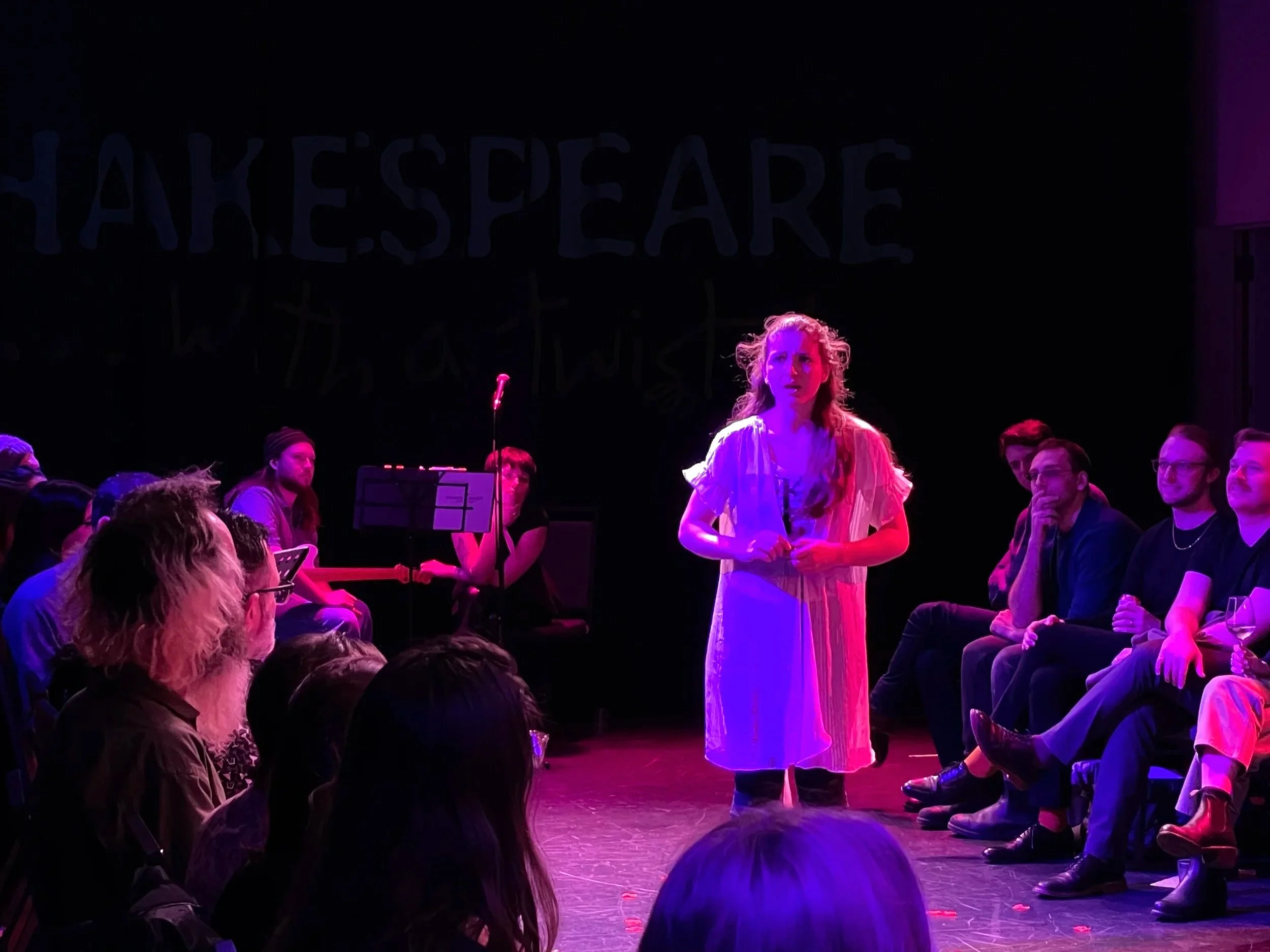 A woman standing on stage under purple and pink lighting, surrounded by seated audience members in a dark theater.