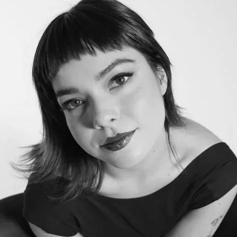 A black and white portrait of a young person in lipstick with short dark hair and bangs, wearing a dark top and smiling slightly.