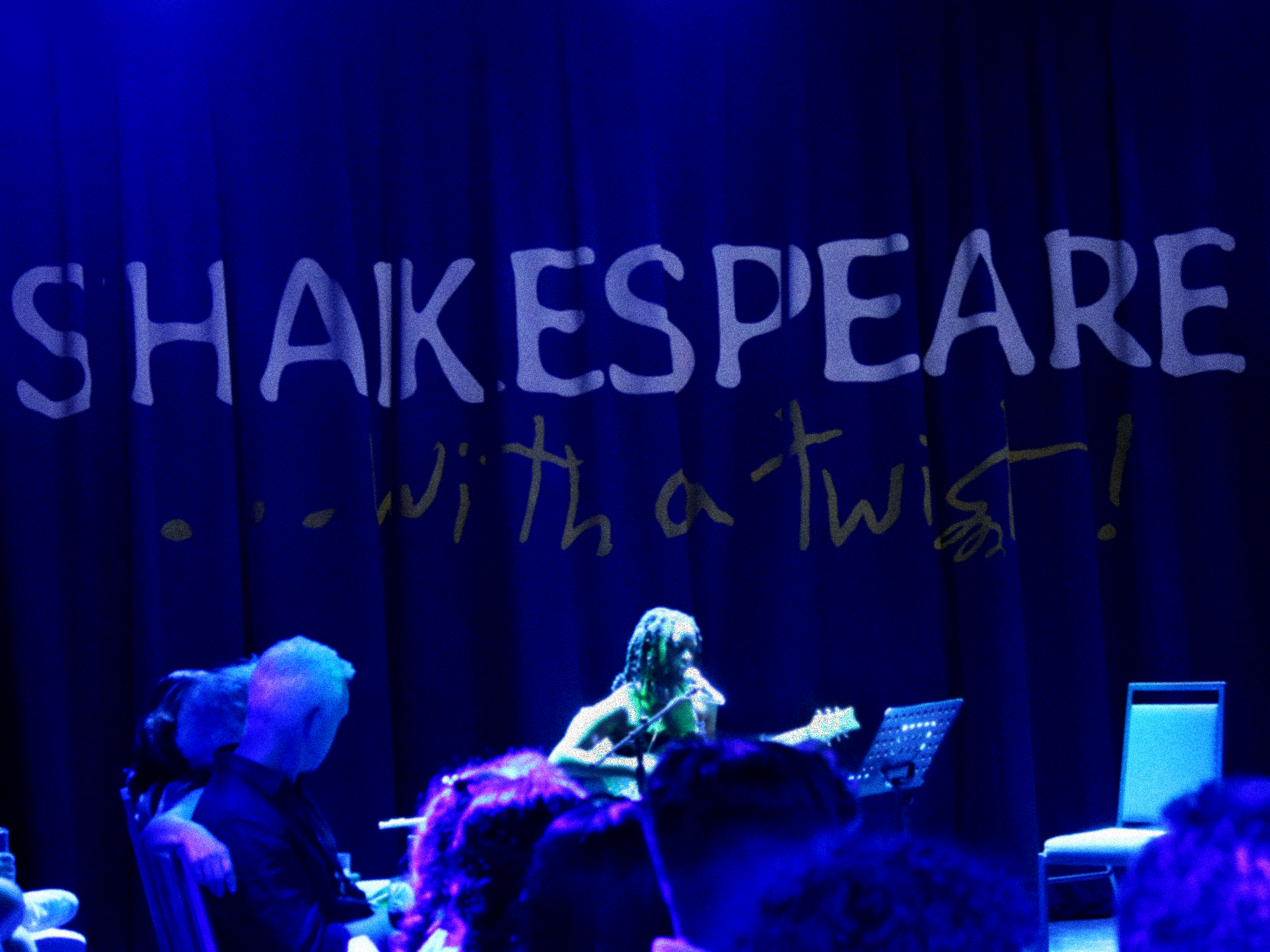 A stage with a blue curtain and a sign that reads 'SHAKESPEARE... with a twist!' in illuminated letters. A musician is seated in front of the curtain with music stands and sheet music.