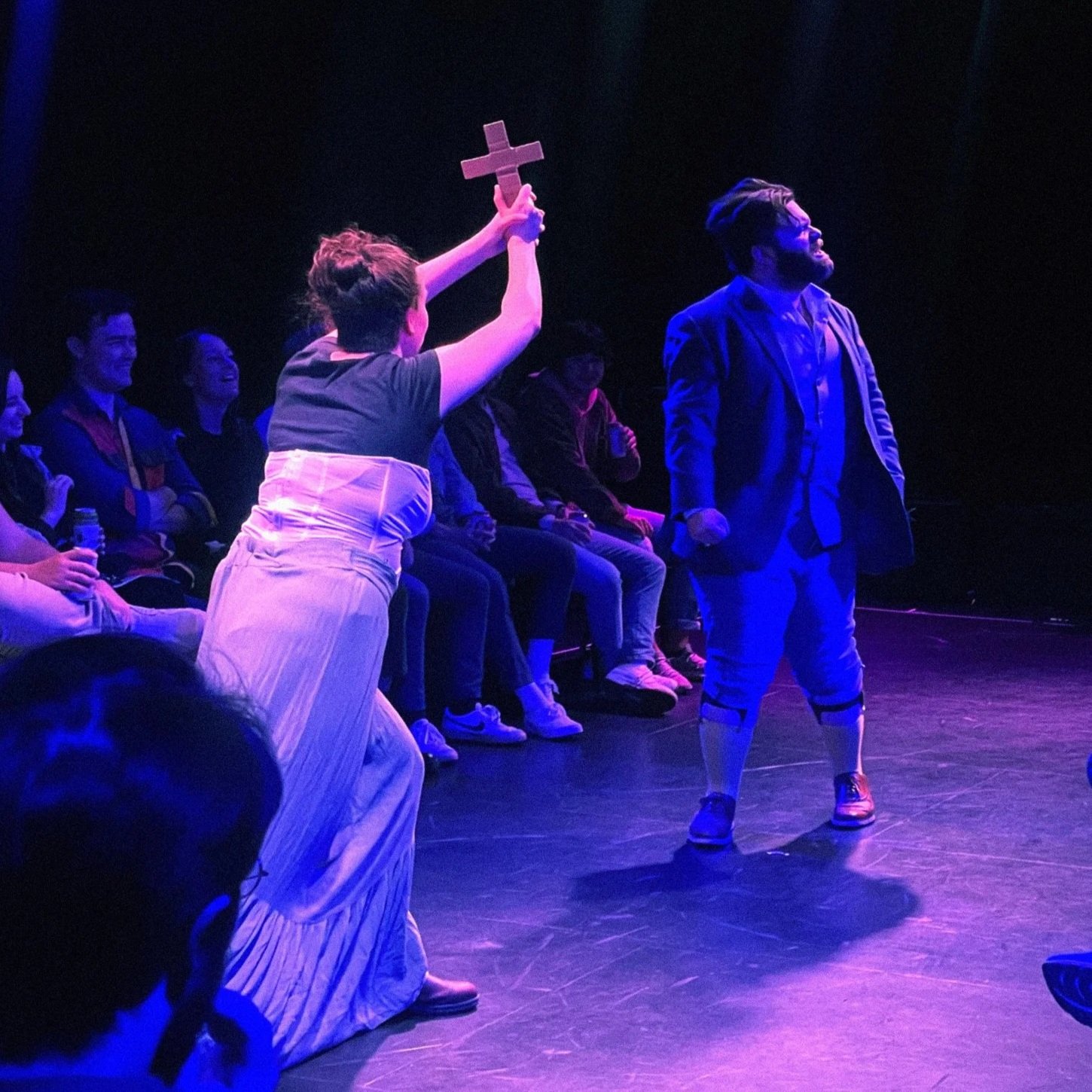 A theatrical performance with a person holding a cross prop and a man dressed in a suit jacket and yellow socks on stage, with an audience watching in the background.