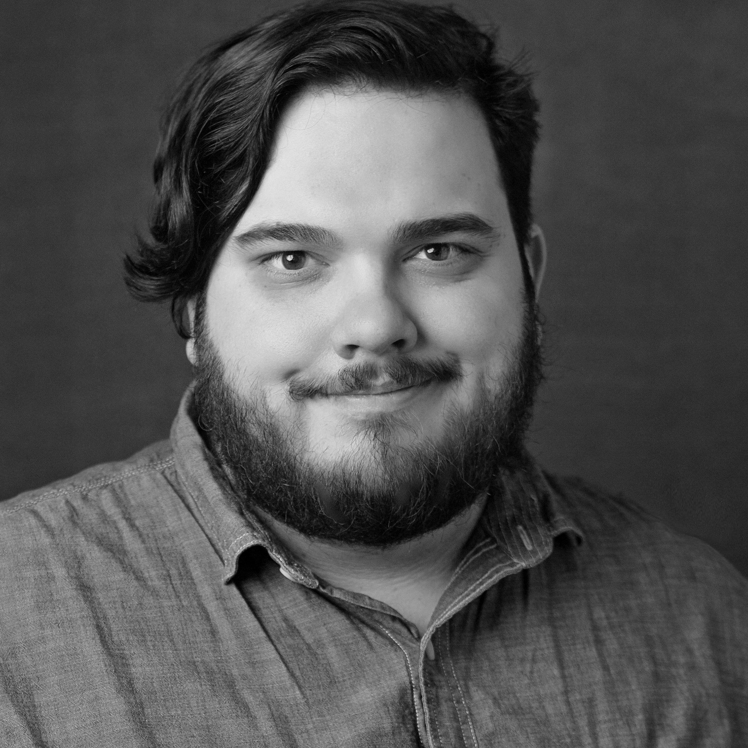 Close-up black-and-white portrait of a young man with wavy hair, beard, wearing a collared shirt.
