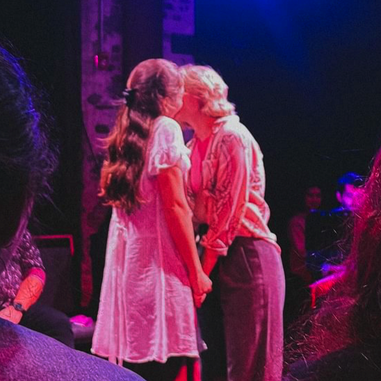 Two women are standing close and leaning in for a kiss on stage, surrounded by an audience in a dimly lit setting with colorful lights.