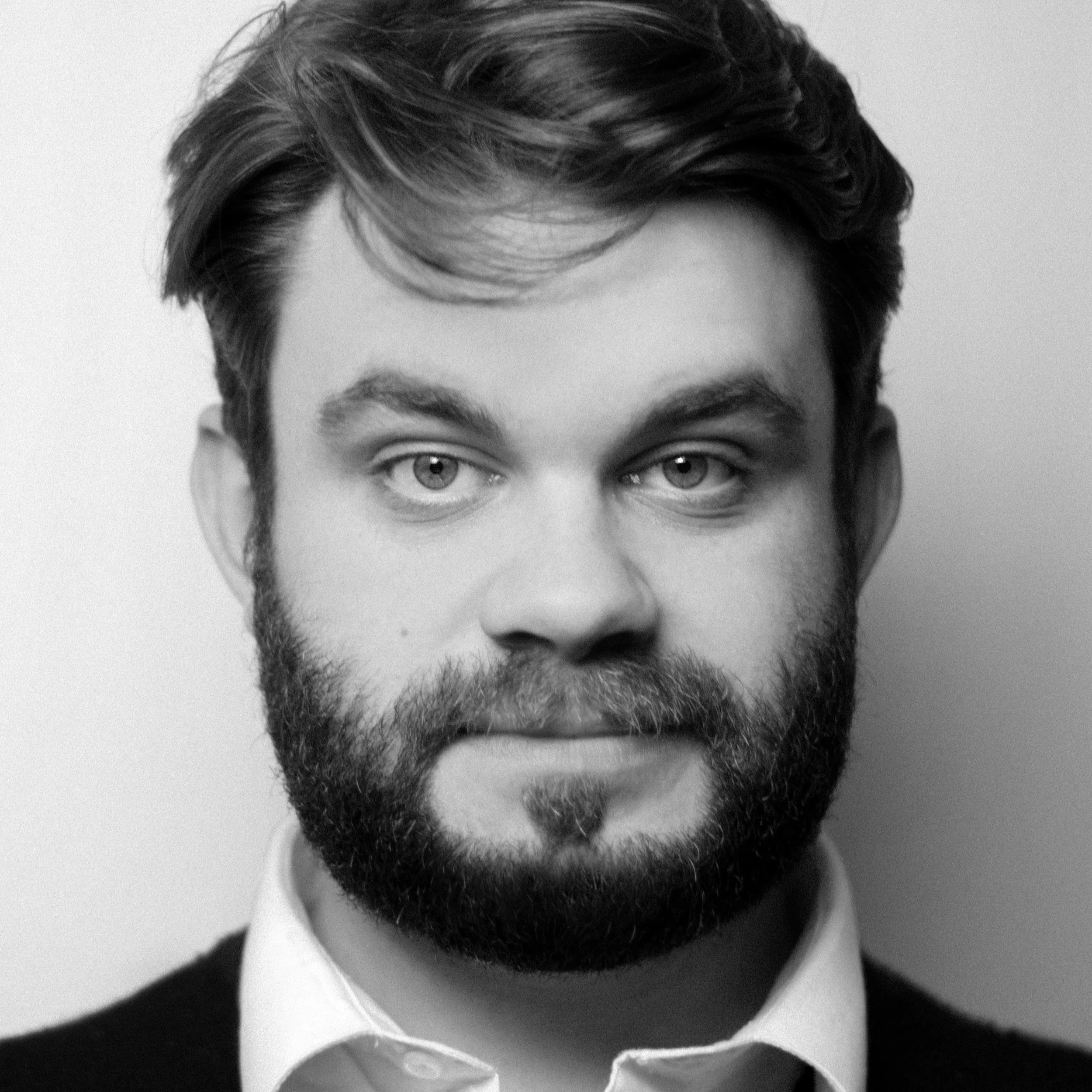 A black and white close-up of a man's face, showing his eyes, eyebrows, nose, and a full beard and mustache. He has dark, wavy hair, and is wearing a collared shirt.