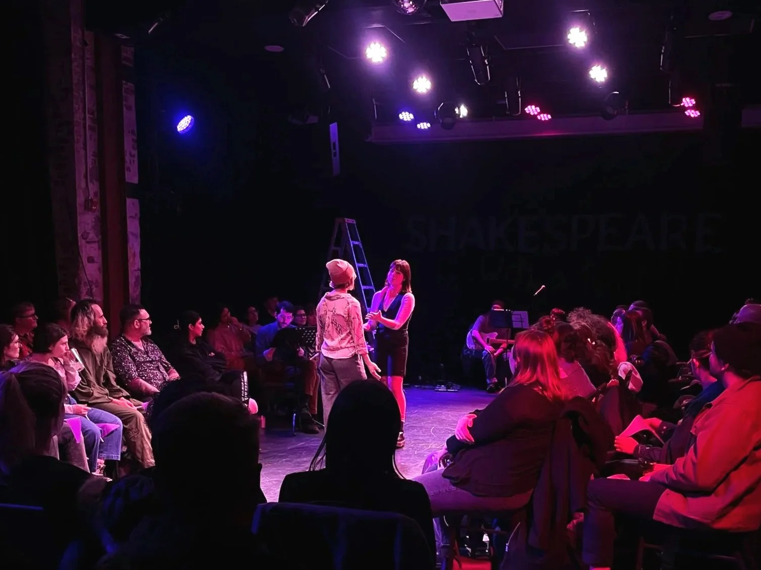A stage performance with an audience watching. Two performers are center stage engaged in dialogue, with stage lights illuminating them. Several audience members are sitting around in a darkened theater with purple lighting.