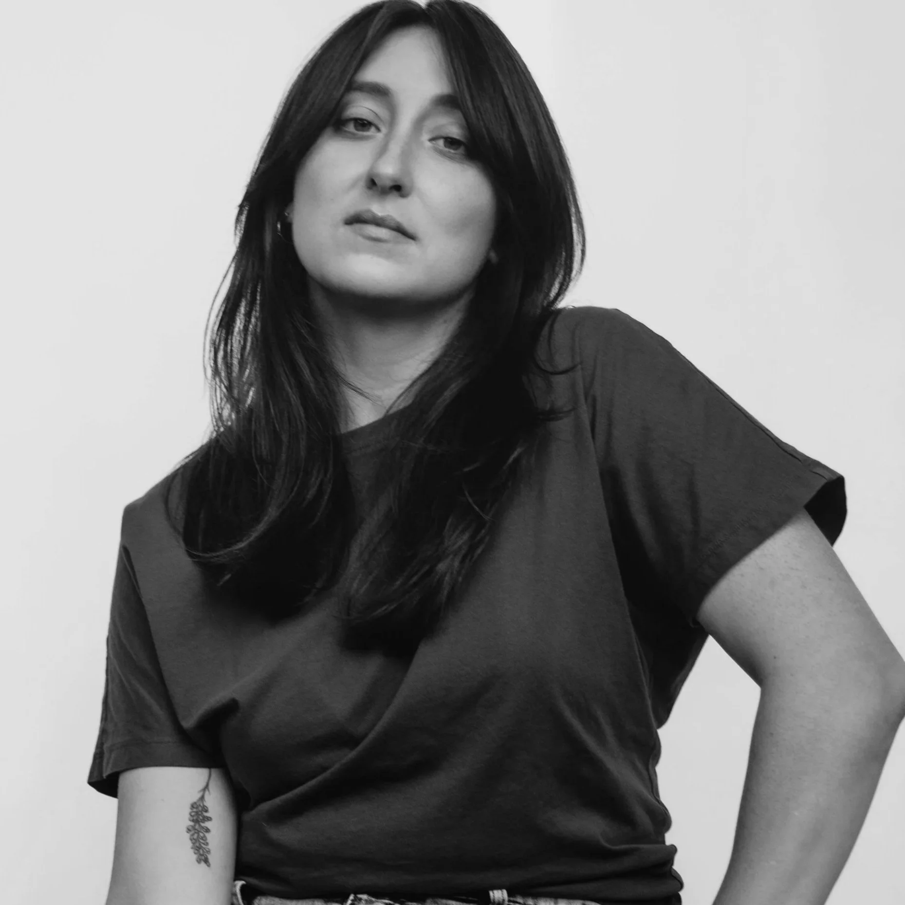 Black and white photo of a woman with long dark hair, wearing a dark t-shirt, standing against a plain light background.
