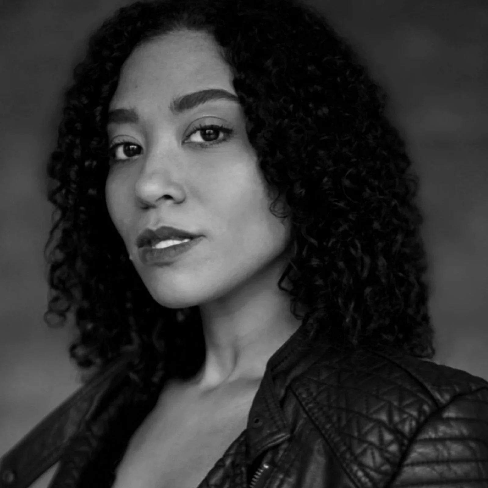Black and white portrait of a person with curly hair, wearing a leather jacket, looking slightly to the side.