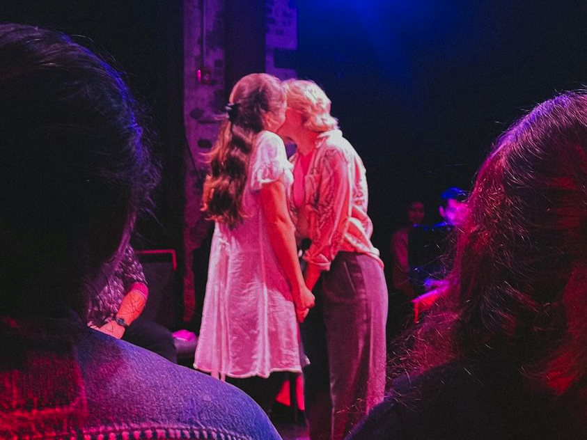 Two women are sharing a kiss on stage, with one in a light dress and the other in a striped shirt and pants, surrounded by an audience and dim stage lighting.