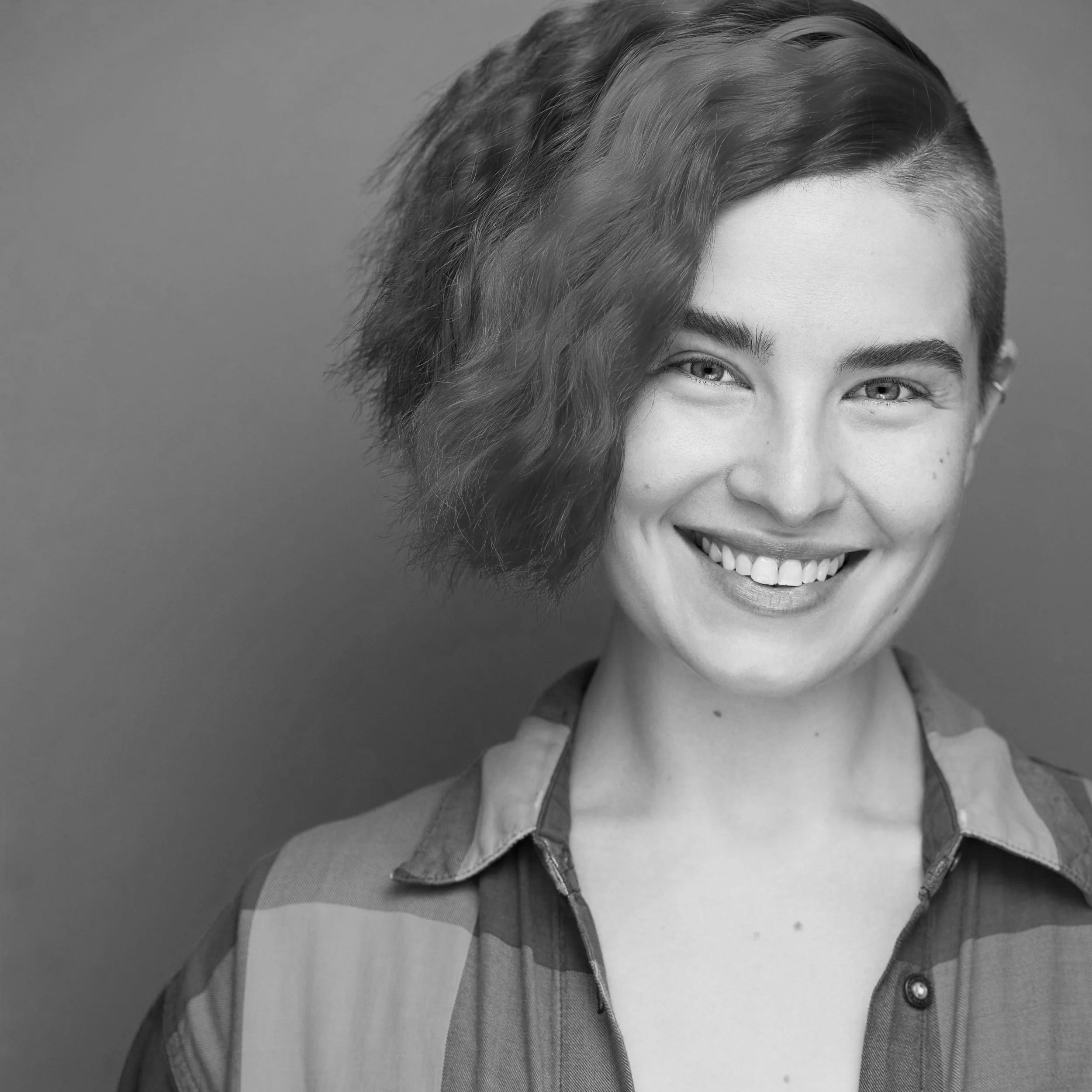 Close-up black and white portrait of a person smiling, showing their teeth. They have short hair styled with waves, and are wearing a collared shirt.