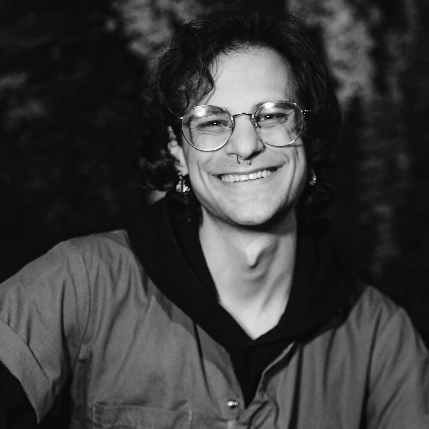 A person with curly hair wearing round glasses and a nose ring, smiling in black and white.