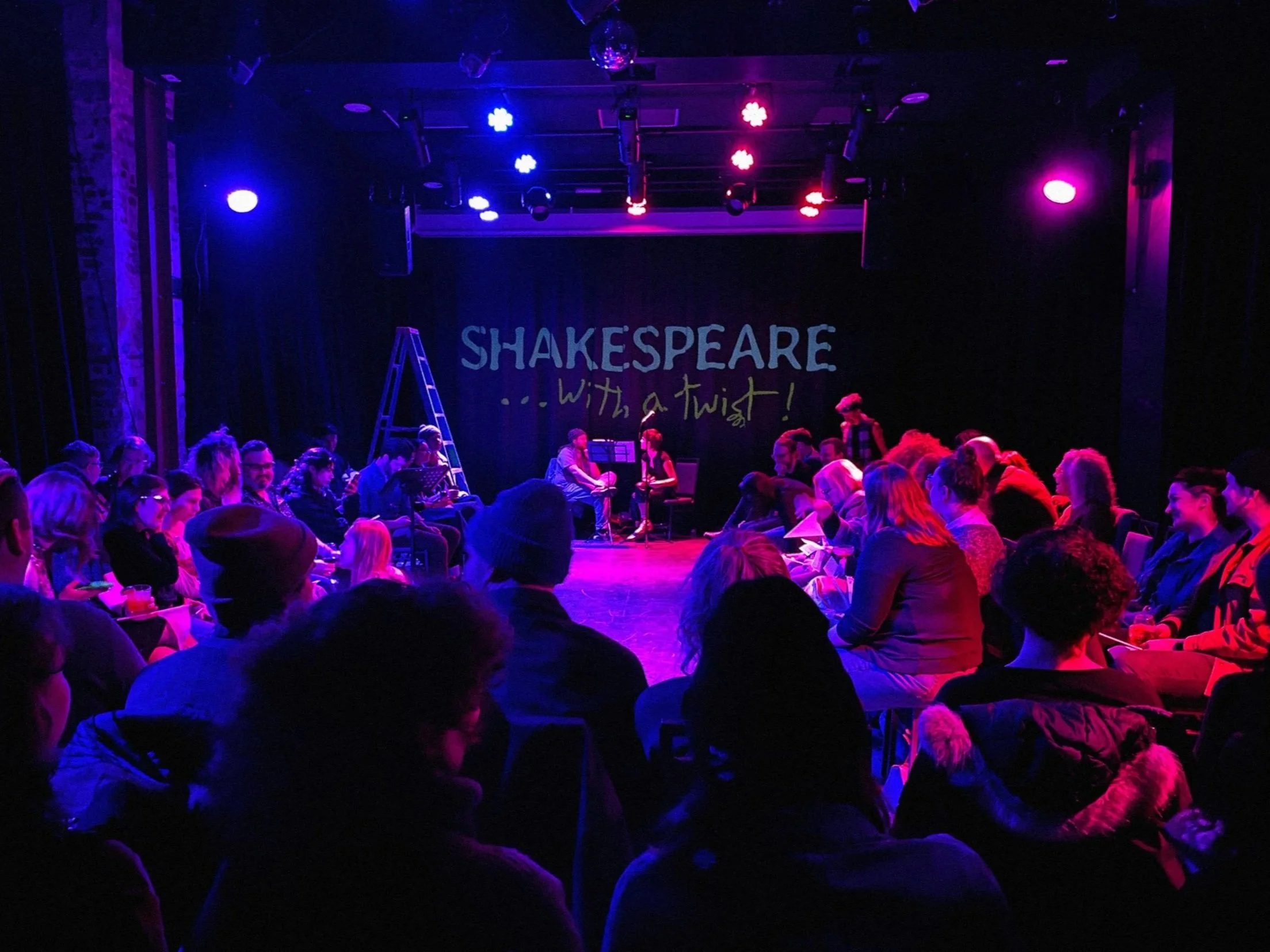 A theater stage with the words "Shakespeare... with a twist!" in the background, illuminated by colorful stage lights, and an audience seated facing the stage.