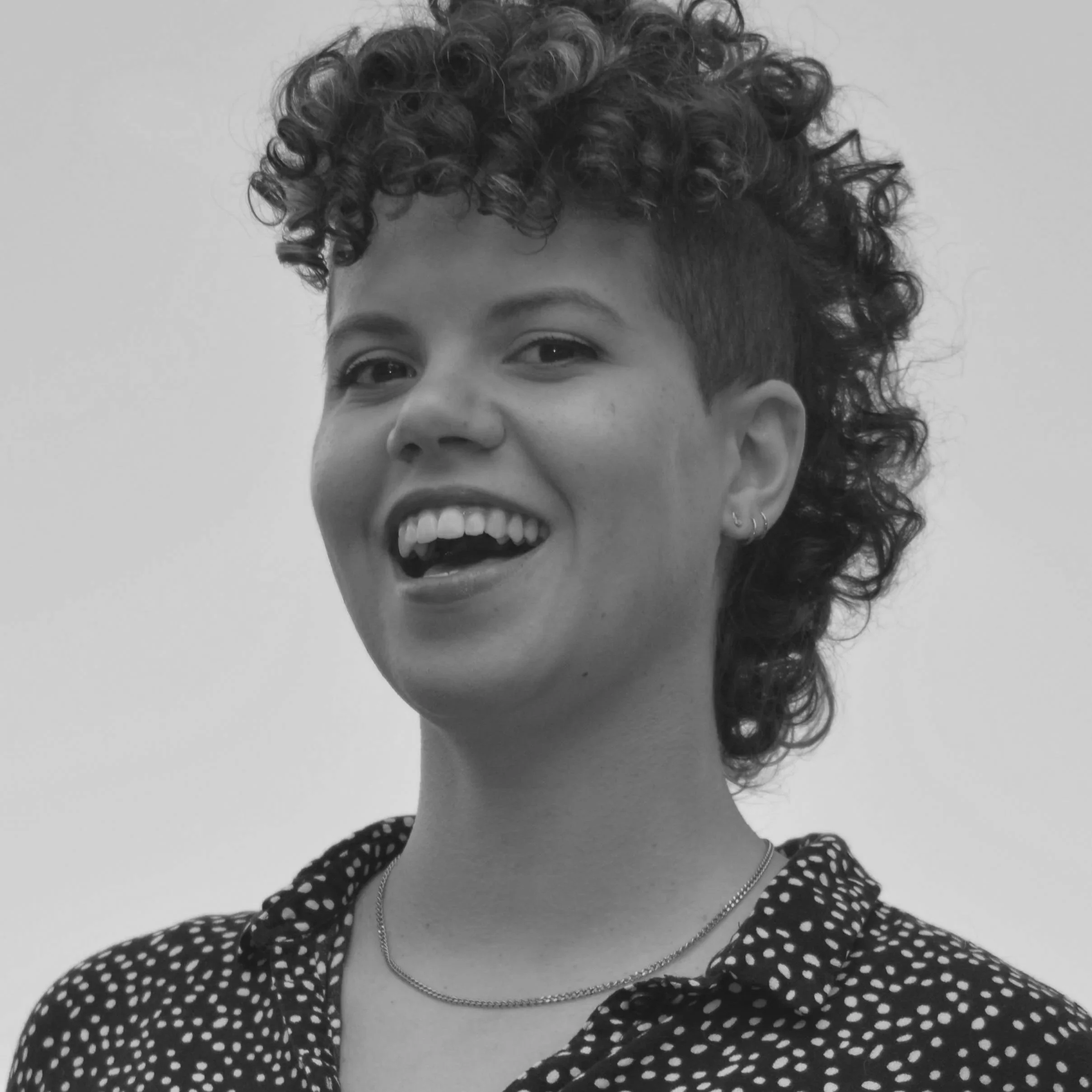 Close-up of a smiling person with curly hair, wearing earrings, a necklace, and a polka dot shirt, against a plain background.