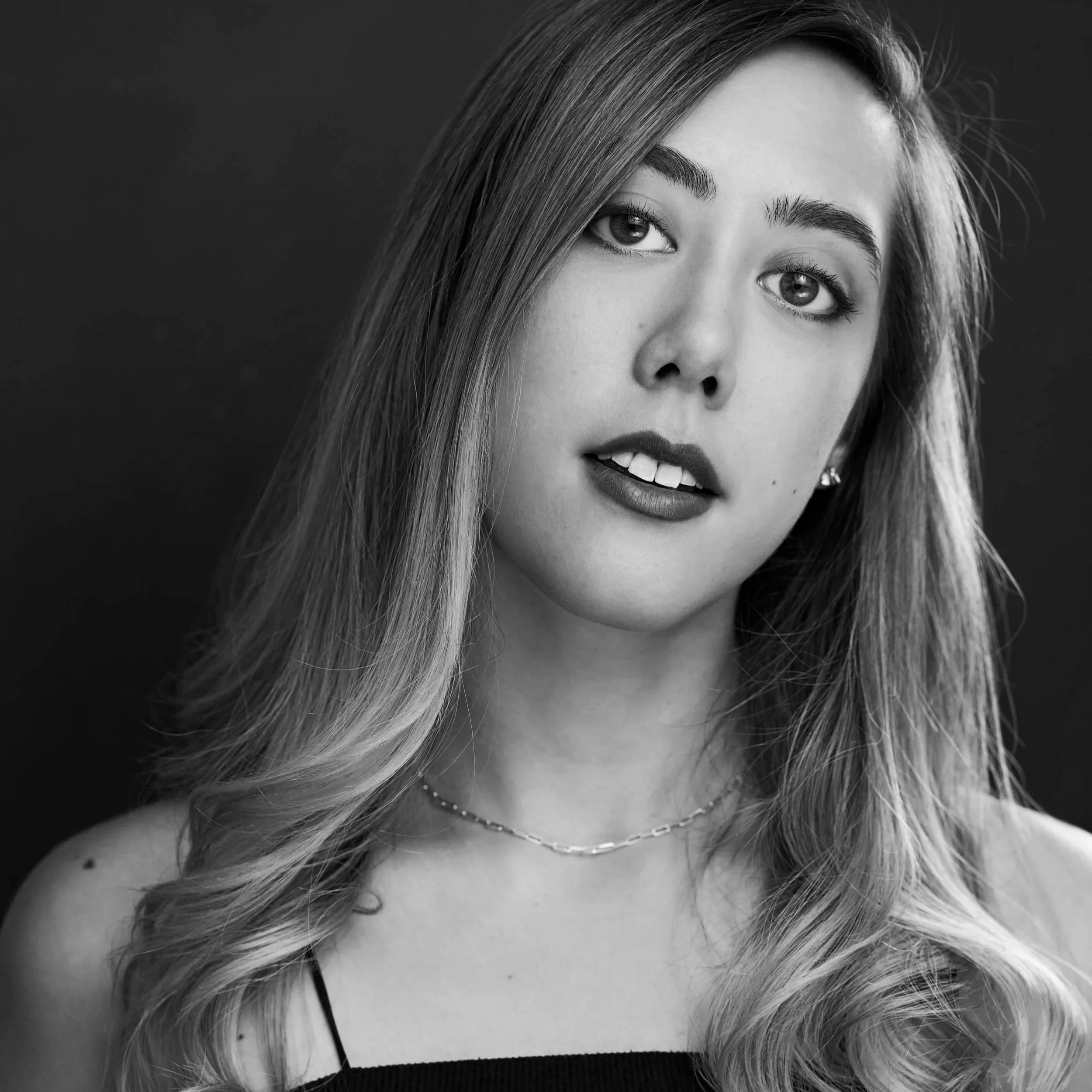 A close-up black and white portrait of a young woman with long hair, looking directly at the camera with a slight smile.
