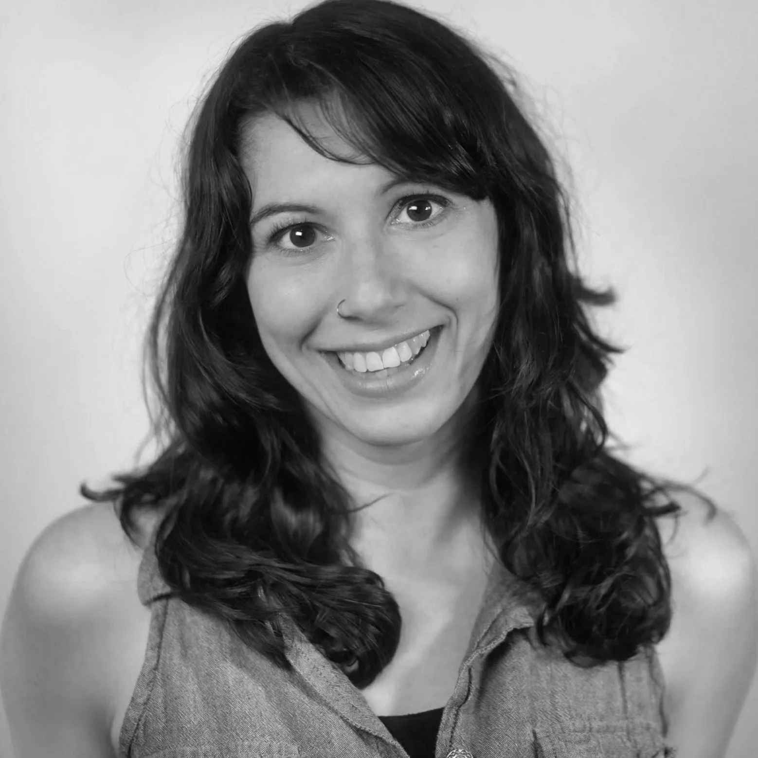 Black and white photo of a woman with wavy dark hair, smile, and nose ring, wearing a shirt with collar.