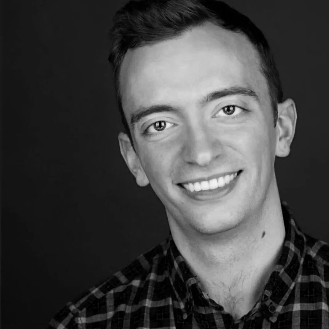 Black and white portrait of a young man smiling, with short dark hair, wearing a checkered shirt, against a dark background.