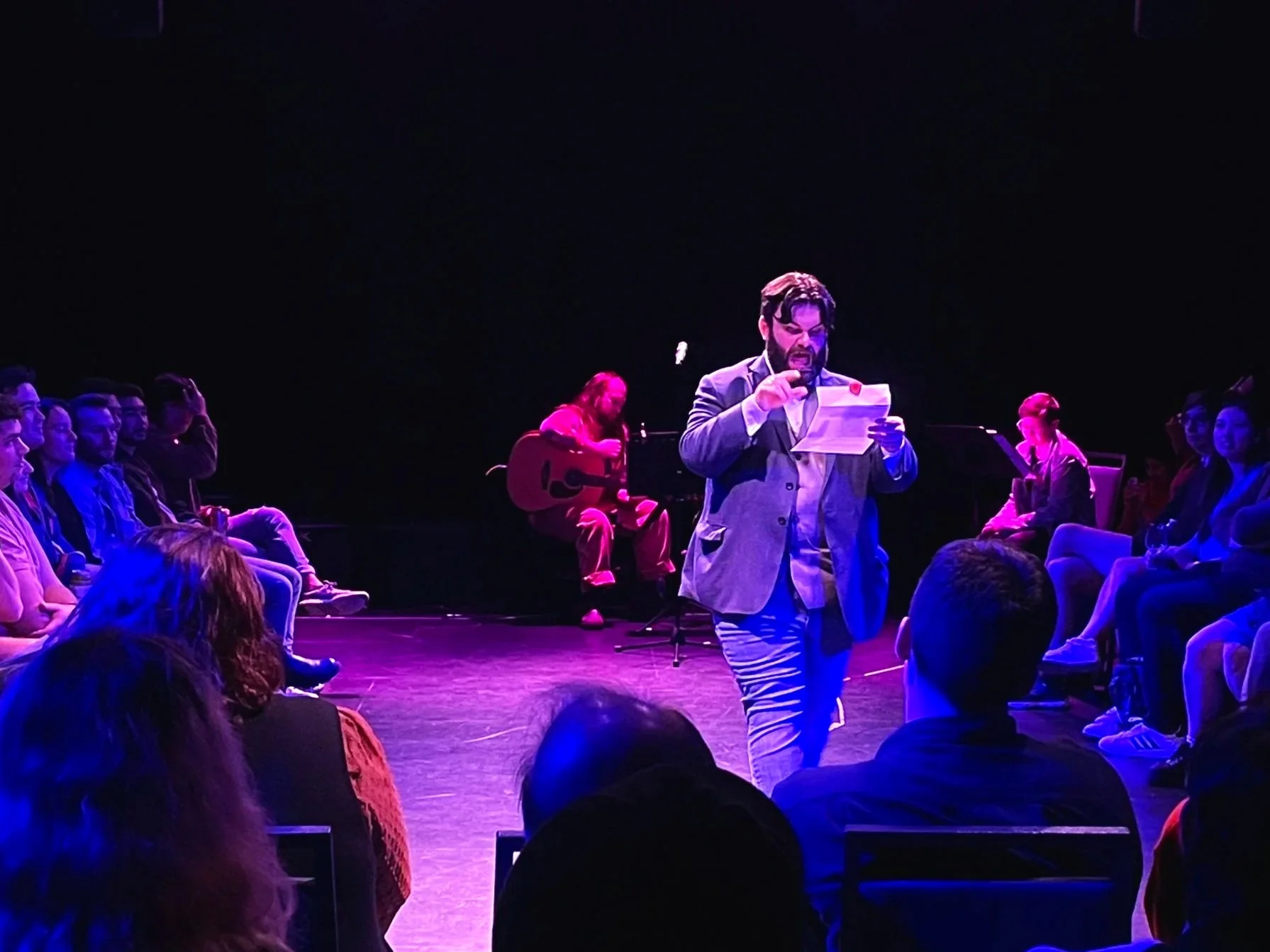 A man in a gray blazer standing on stage, reading from a piece of paper, in front of a seated audience. In the background, a person plays a guitar and others are seated, watching the performance. The stage is lit with purple and blue lights.