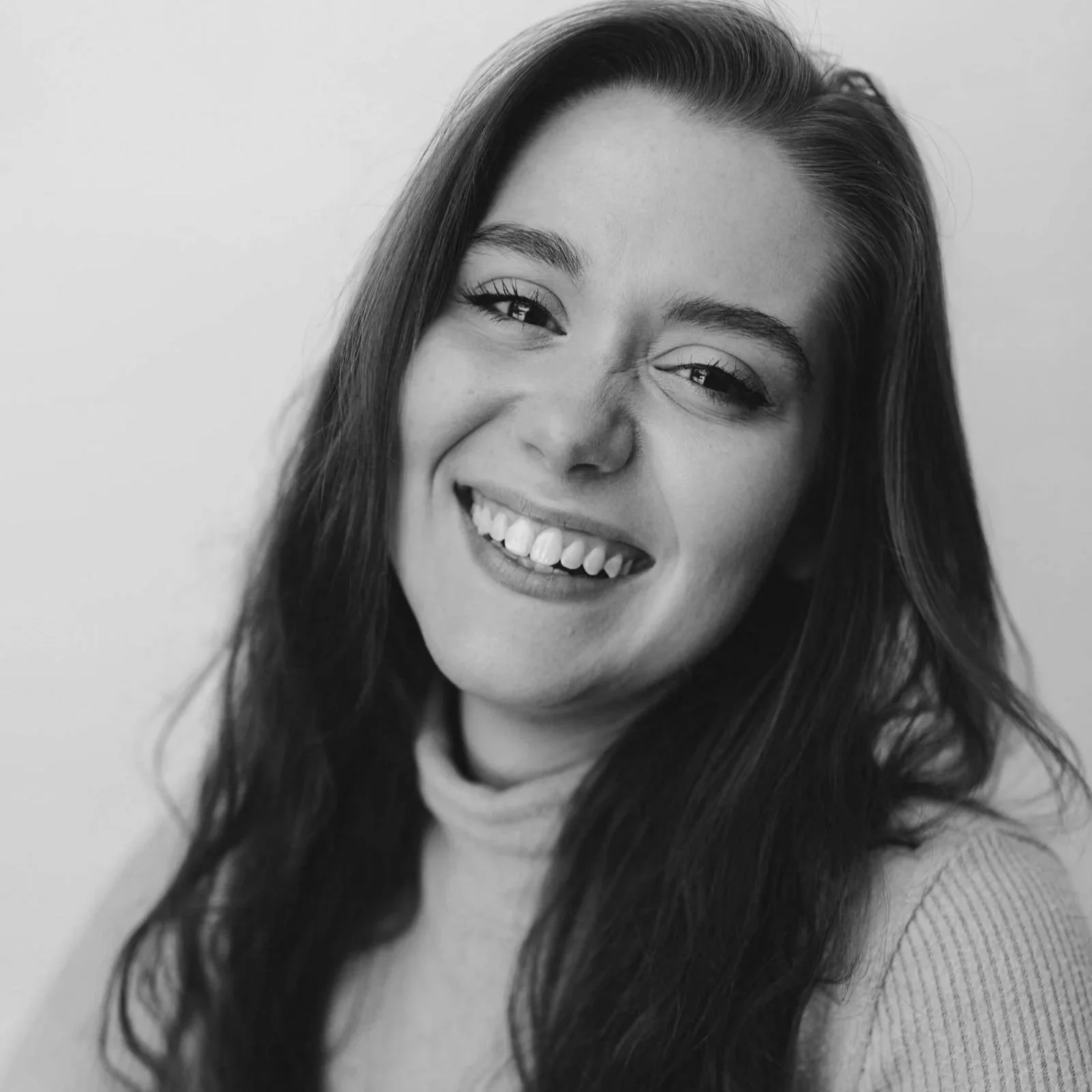 Black and white close-up portrait of a young woman smiling, showing her teeth, with long dark hair and expressive eyes.