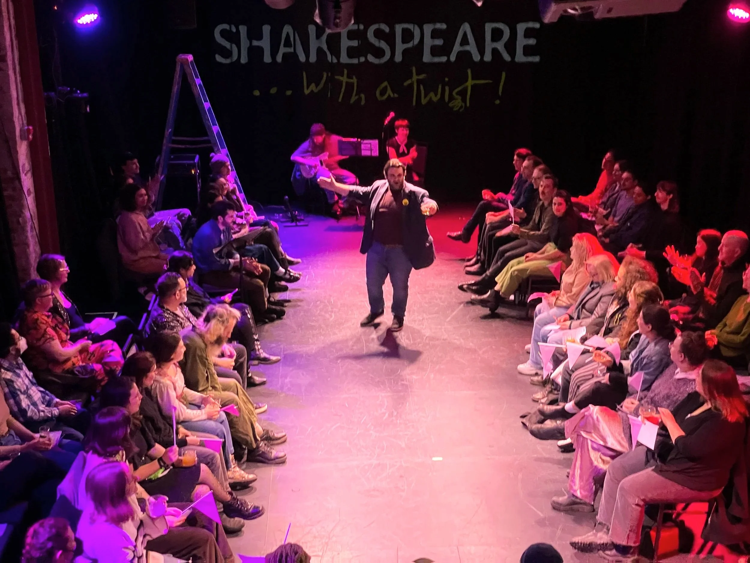 Audience seated on both sides of a stage with a man walking in front of the audience holding a drink.