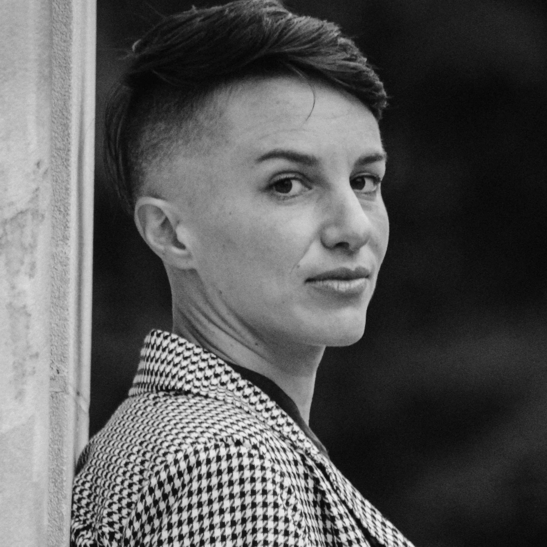 Close-up black and white photo of a person's face with short, styled hair, looking over their shoulder, with a patterned jacket, positioned near a wall.
