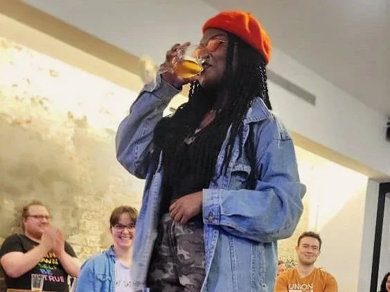 A woman wearing a red beret, red glasses, and denim jacket is drinking from a beer glass.
