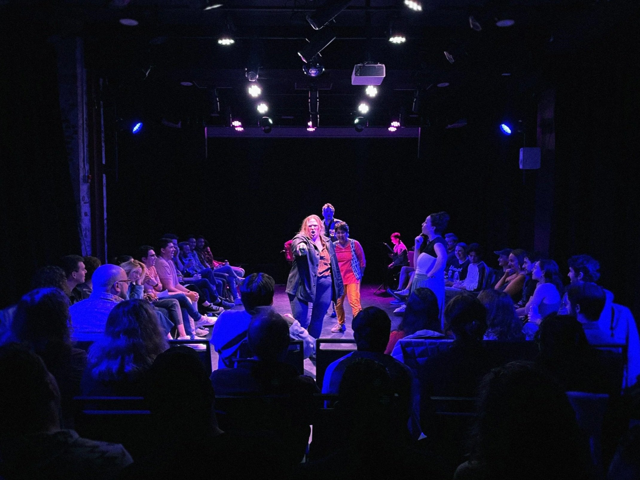 A group of people sitting in a dark room with stage lighting, some of them are smiling and looking toward the front. One actor is standing and pointing forward.