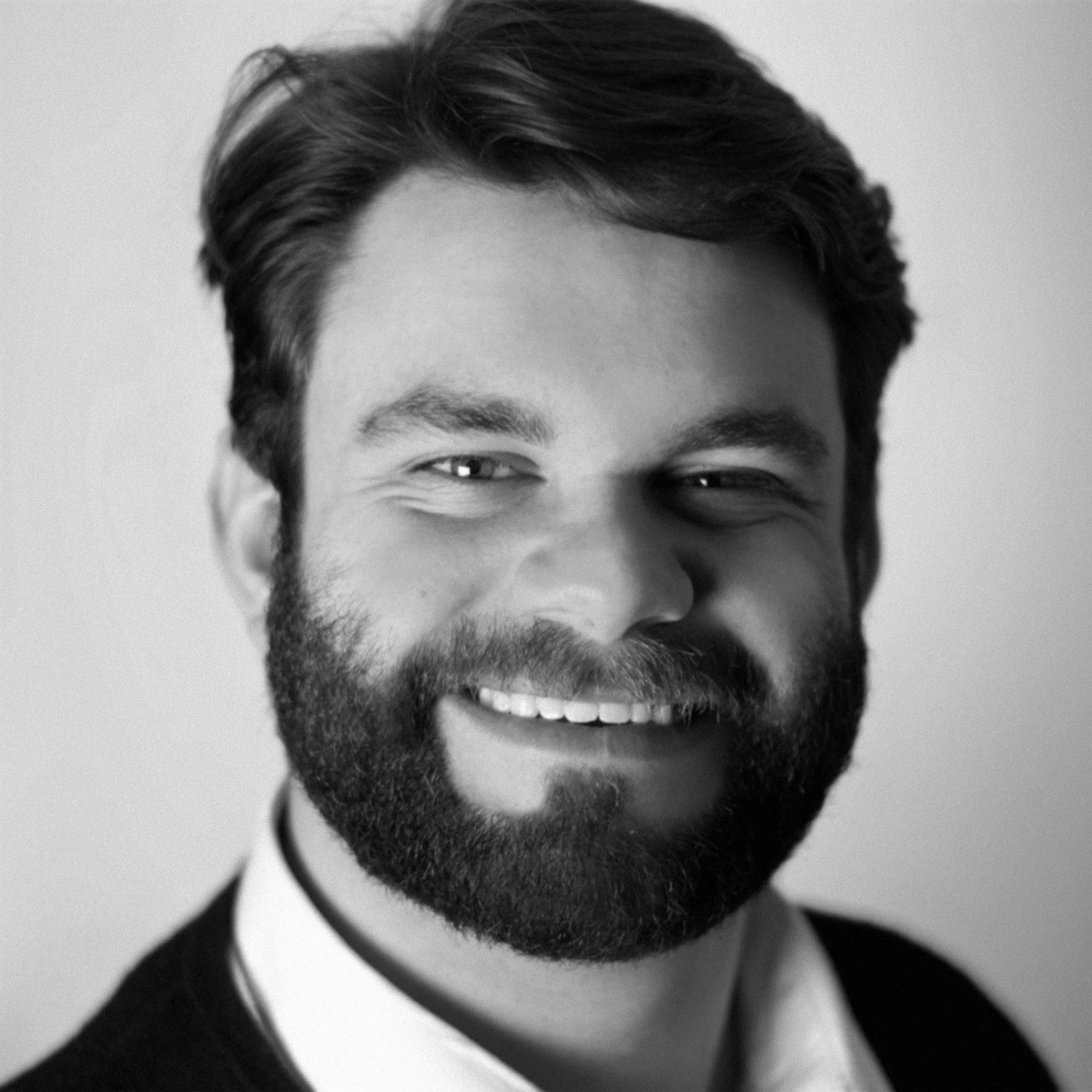 A black-and-white close-up photograph of a man's face with a full beard, smiling, wearing a white collared shirt.
