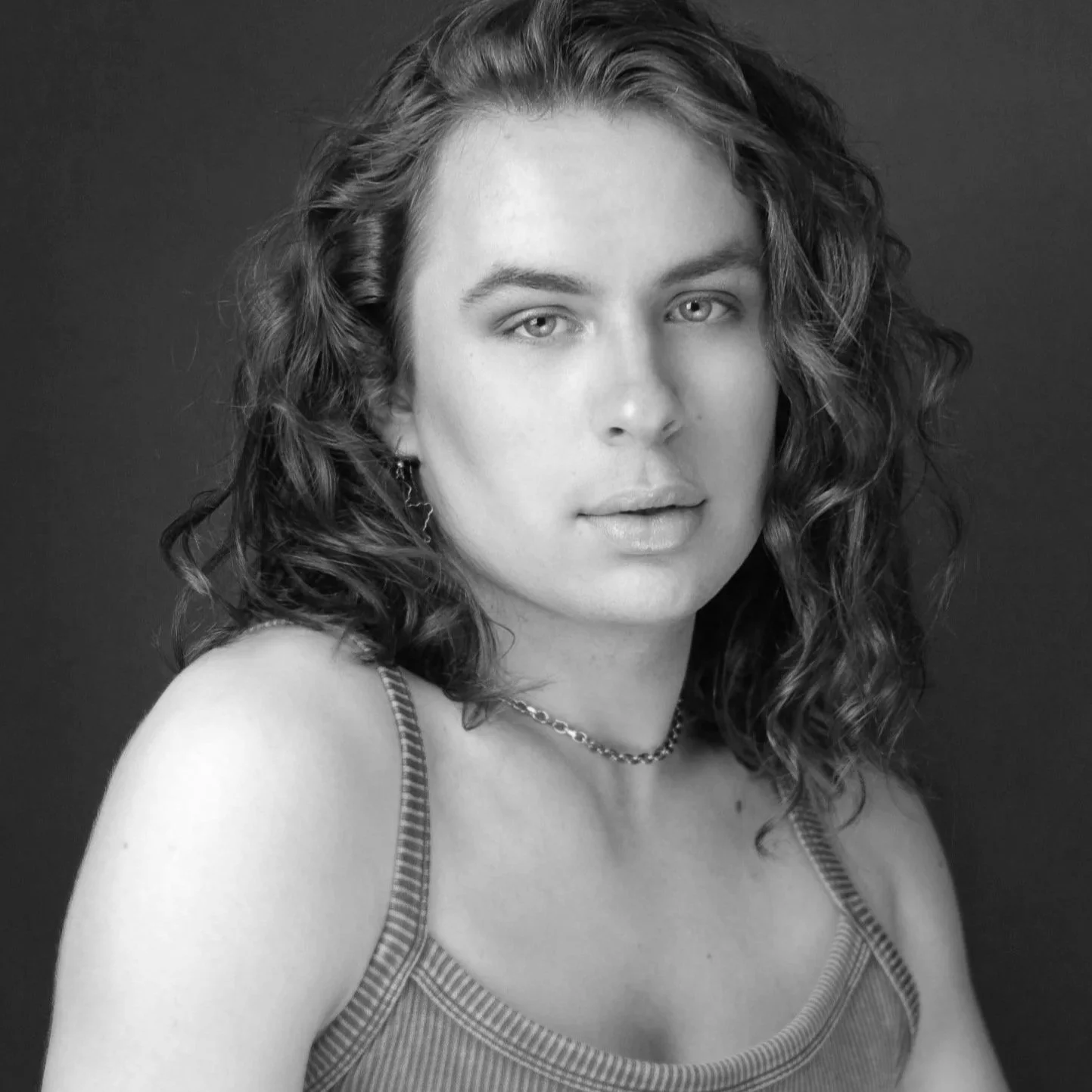 Black and white portrait of a person with long curly hair, wearing a sleeveless top with thin straps, a chain necklace, looking directly at the camera with a neutral expression.