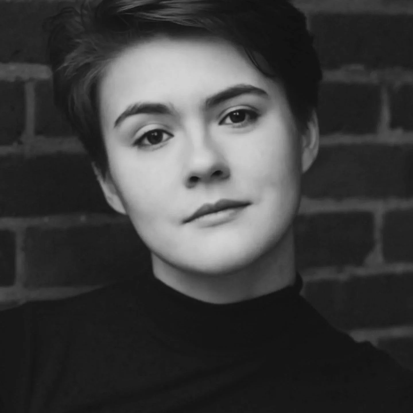 Black and white portrait of a person with short, wavy hair, looking directly at the camera, against a brick wall background.