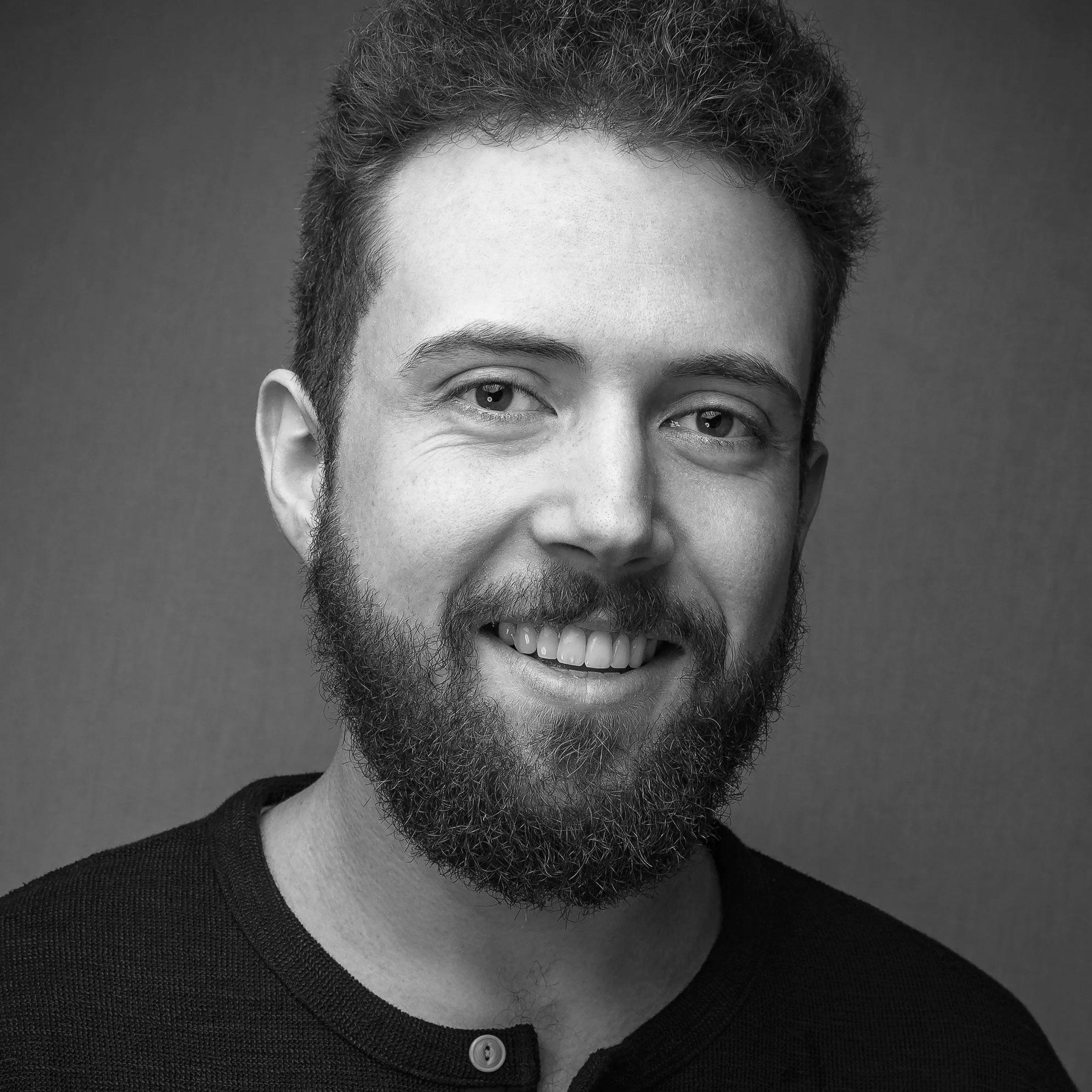 Black and white close-up of a smiling man with hair and beard, wearing a dark textured shirt with a button.