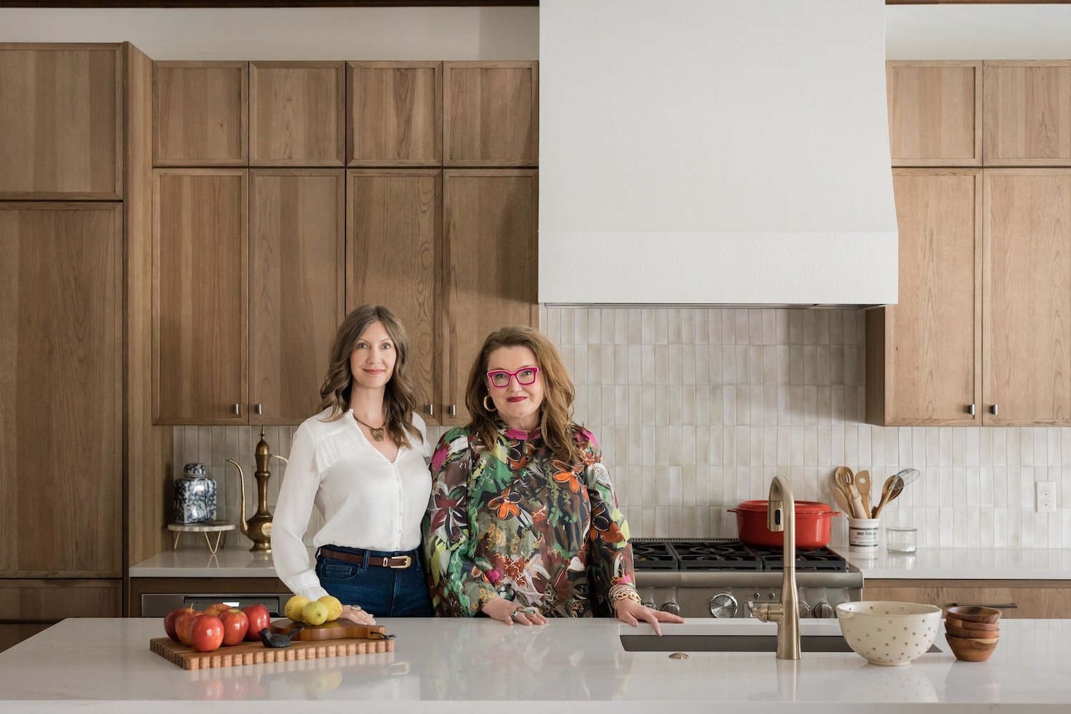 Two women standing in a modern kitchen, one wearing a white blouse and the other in a colorful floral top with pink glasses, with apples on the kitchen counter and kitchen cabinets in the background.
