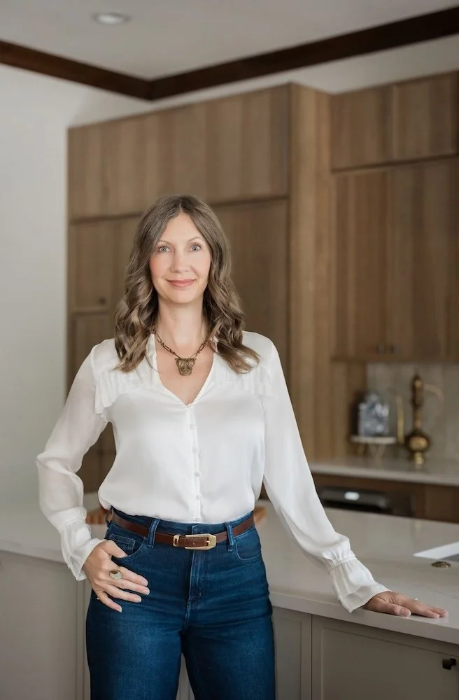 A woman with shoulder-length wavy hair, wearing a white blouse and blue jeans, standing in a modern kitchen with wooden cabinets.
