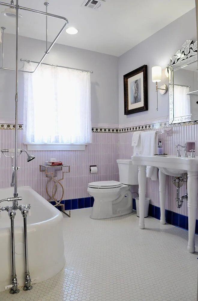 A vintage-style bathroom with a white bathtub, a toilet, a pedestal sink, and a large mirror. Light-colored walls with decorative border tiles, a framed picture, and a small side table by the window with sheer curtains.