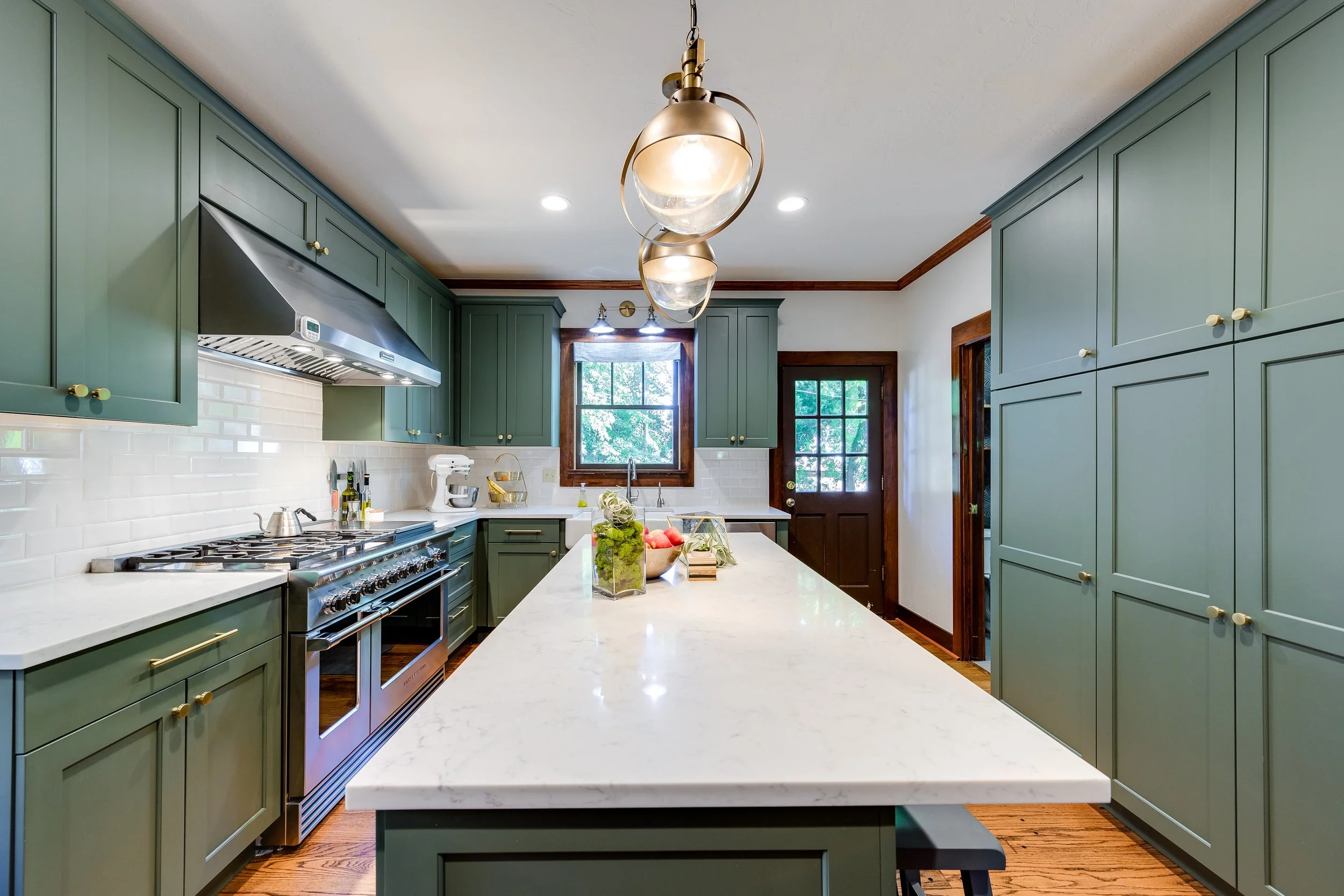 A modern kitchen with green cabinets, white countertops, and a central island. There is a stainless steel stove, a white backsplash, and a window with greenery outside. Pendant lights hang from the ceiling, and a door with glass panes leads outside.
