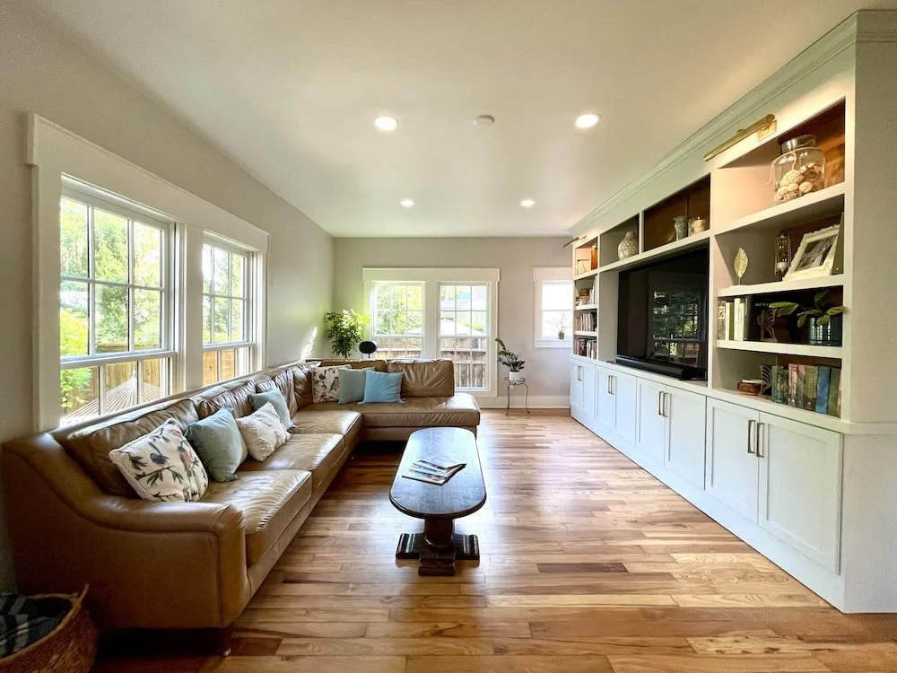 Living room with large windows, a tan sectional sofa with pillows, a wooden coffee table, and a white built-in entertainment center with a TV and decorative items.
